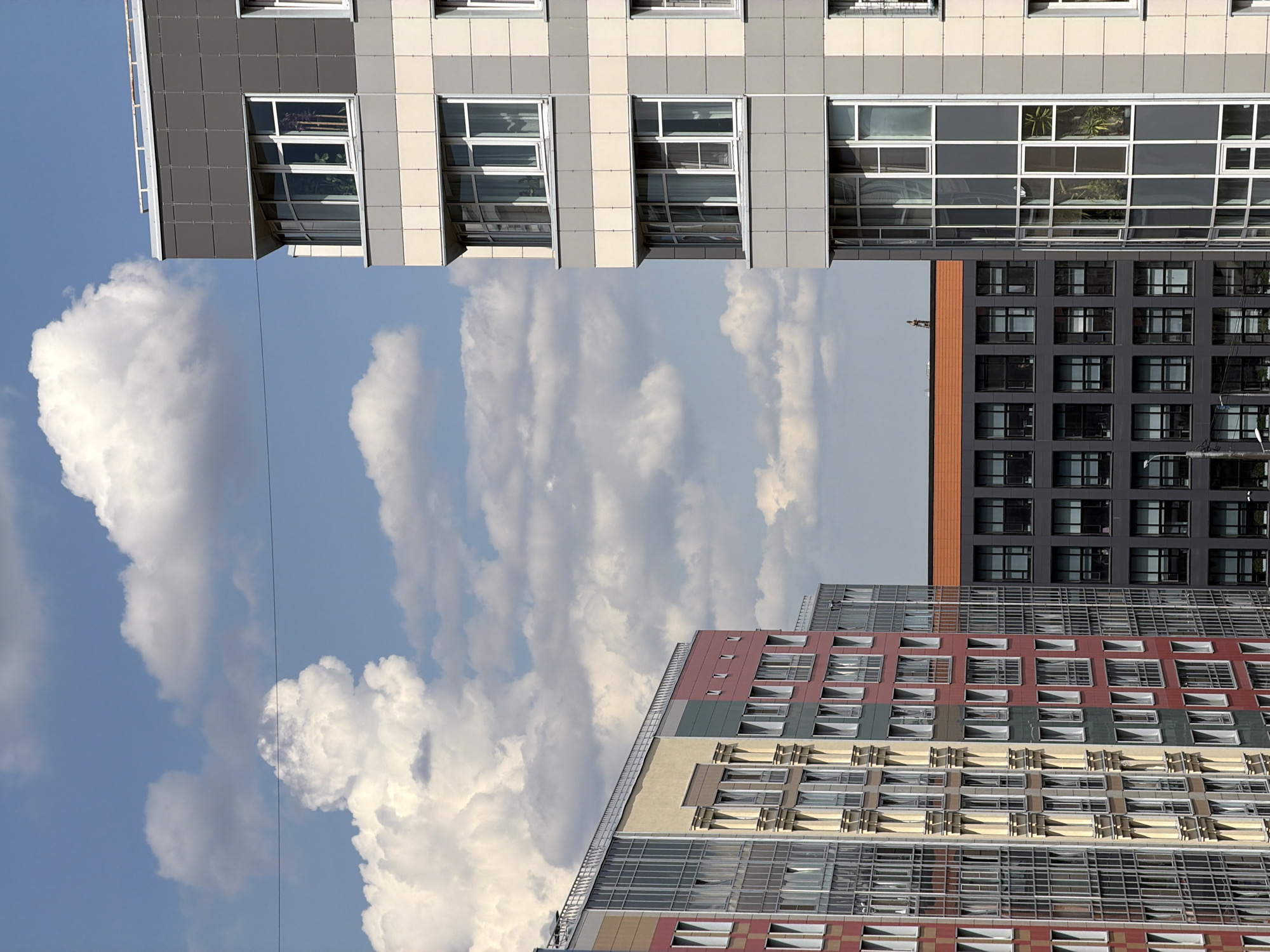 This image captures a vertical urban landscape featuring multiple modern high-rise buildings against a bright, cloudy sky. The overall scene suggests a contemporary city environment during daylight hours.
The sky, occupying a significant portion of the image, is a clear blue adorned with numerous large, white, fluffy cumulus clouds. A thin, dark wire or power line stretches across the sky, running horizontally through the middle of the frame.
Three distinct modern buildings are visible. In the upper left part of the image, a light-colored building, primarily white and light gray, stands tall. Its facade is composed of rectangular panels and features large, multi-pane windows, some of which are reflective, mirroring the sky and clouds. Interior views through some windows reveal plants.
Below and to the right of this light-colored structure, two other buildings are present. One building, located in the mid-right section, is characterized by its dark gray or black facade, densely packed with square-shaped windows, creating a grid-like pattern. It has a distinctive reddish-brown band along its top edge.
Further to the right and lower in the frame, another multi-story building displays a more colorful facade, featuring panels in shades of red, yellow, and gray. This building also has numerous windows, some of which show varied designs and potentially small balconies.
The architecture of all buildings is modern, characterized by extensive use of glass, geometric patterns, and varied facade materials. There are no visible people, specific activities, or discernible text. The scene is static, showcasing urban development under fair weather conditions.