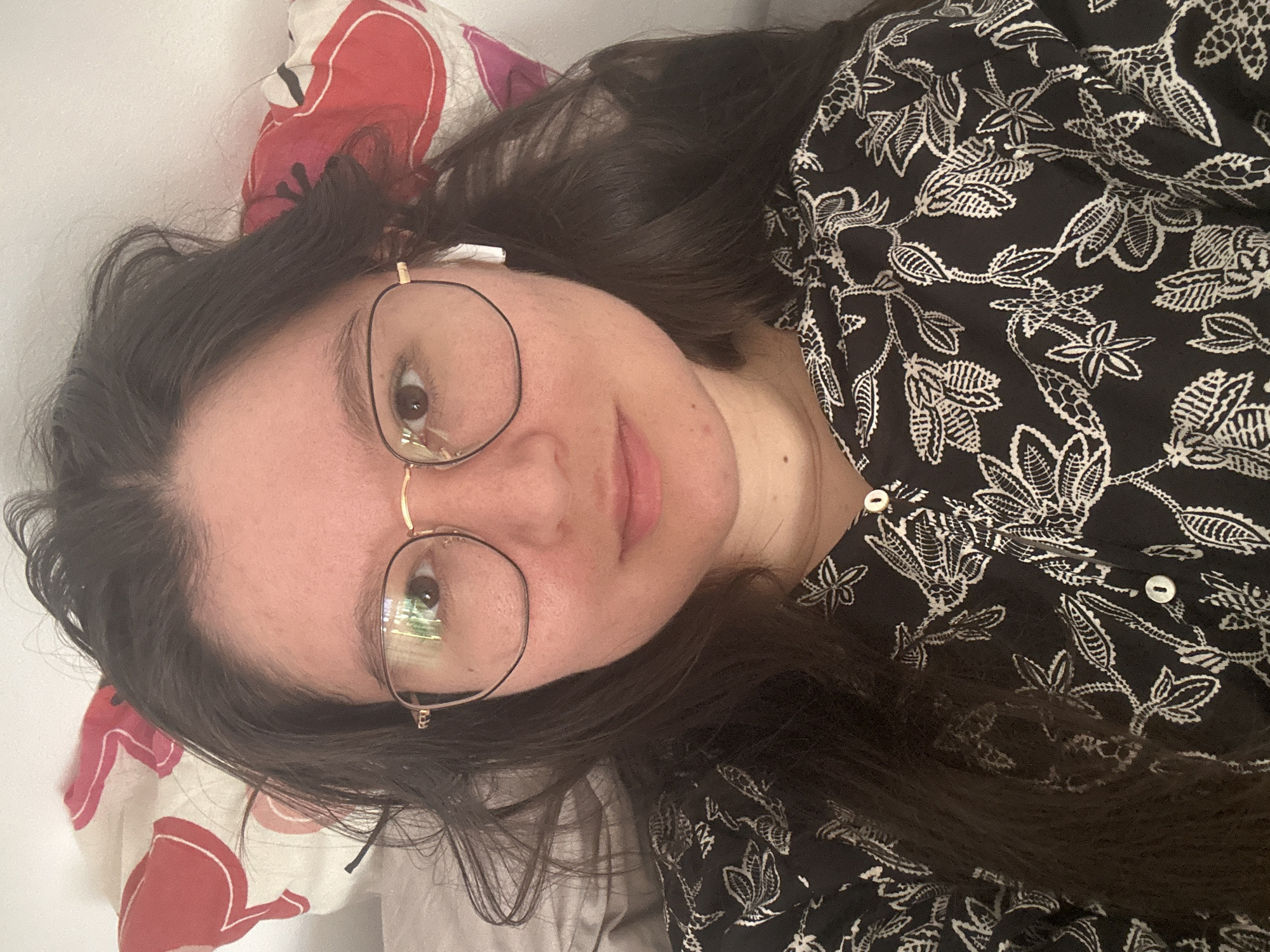 A person is lying on a bed, with a patterned pillow. The person is wearing round glasses and an earbud. The person is also wearing a black top with a white floral design. The scene does not obviously take place in Câmara de Lobos, Portugal.