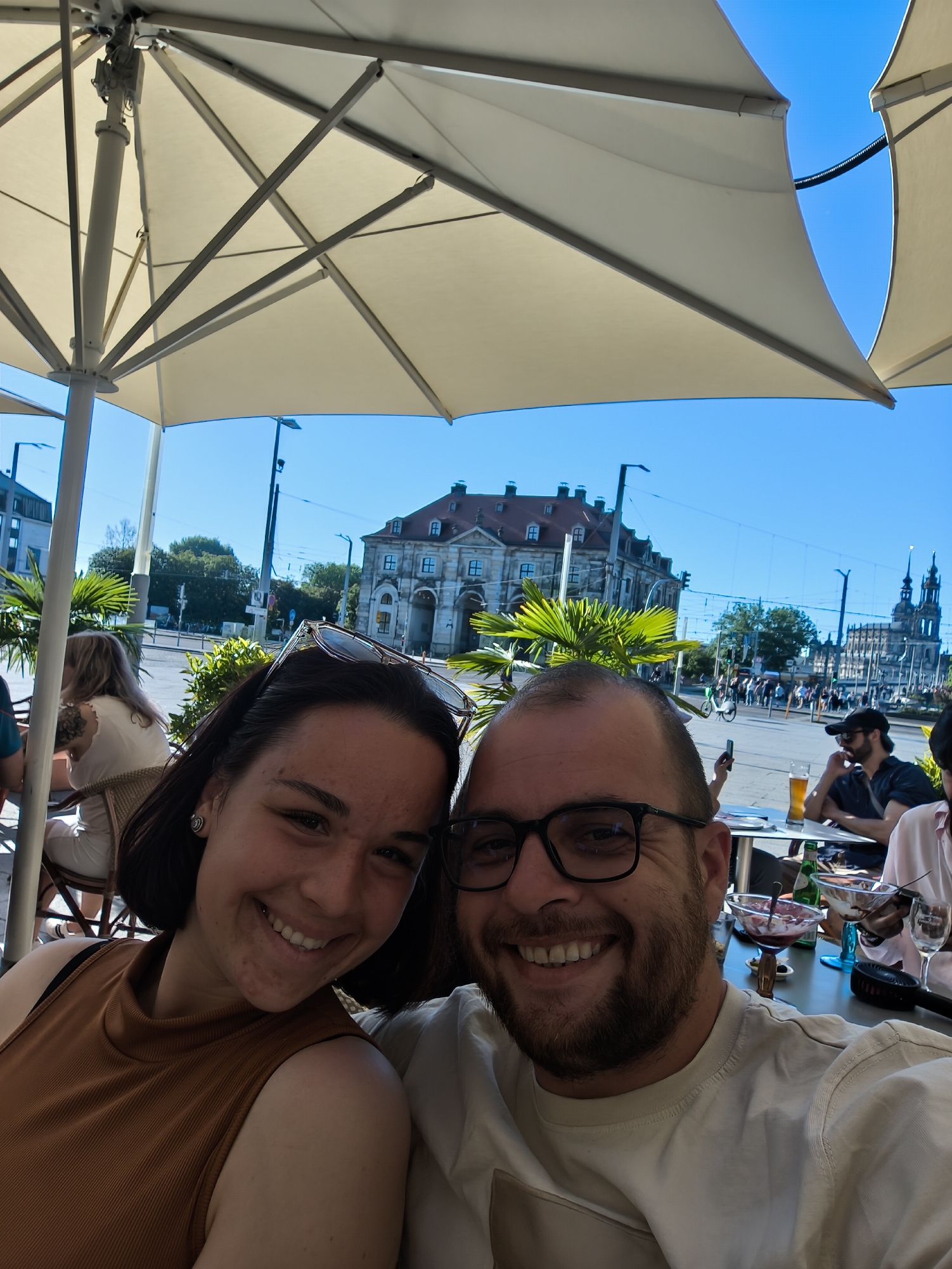 A selfie captures a smiling man and woman seated at an outdoor cafe. The woman is on the left, wearing a brown tank top and sunglasses perched atop her head. The man on the right has a beard and wears glasses and a white shirt. Both are looking at the camera.
Above them, a large white sun umbrella provides shade. In the background, a clear blue sky highlights a city landscape, with historical buildings and what appears to be a church spire visible. Other patrons are seated at nearby tables. Food and drinks are visible on the table in front of the couple. The photograph appears to be taken in Dresden, Germany.