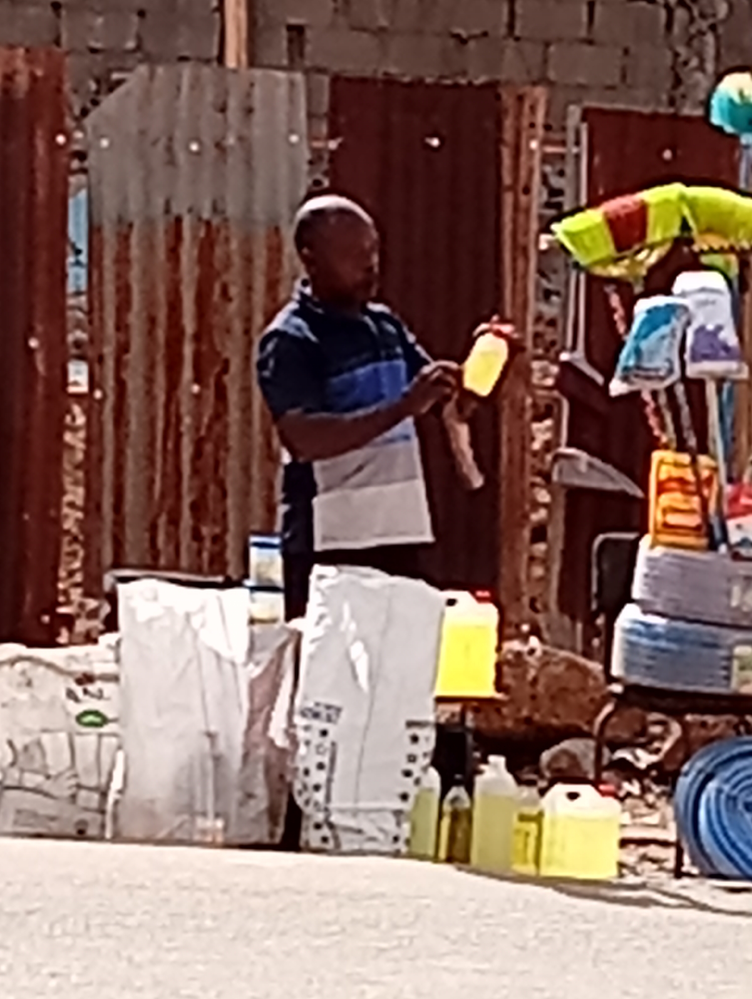 A street vendor operates an outdoor stall, likely in Maiburiji, Nigeria, under bright daylight. The main subject is a dark-skinned man with short or bald hair, wearing a short-sleeved polo shirt with horizontal blue and white stripes. He is holding a small, yellow plastic bottle, appearing to inspect it or arrange his goods.
The stall is set against a backdrop of rusty, corrugated metal sheets, characteristic of informal market structures, with a rough concrete block wall visible behind. The man's merchandise includes several yellow plastic bottles or containers on the ground, likely holding liquids such as cooking oil or detergents. Large white sacks or bags are also part of the display. To the right of the man, various items are hung and stacked vertically, including bright green and yellow brooms or mops, several plastic-packaged goods, and coils of blue hose or rope.
The scene depicts a typical informal market setting, marked by the practical and somewhat makeshift construction of the stall. No specific time of day beyond bright daylight can be determined, nor are there any discernable emotions on the man's face or visible text due to the image quality.