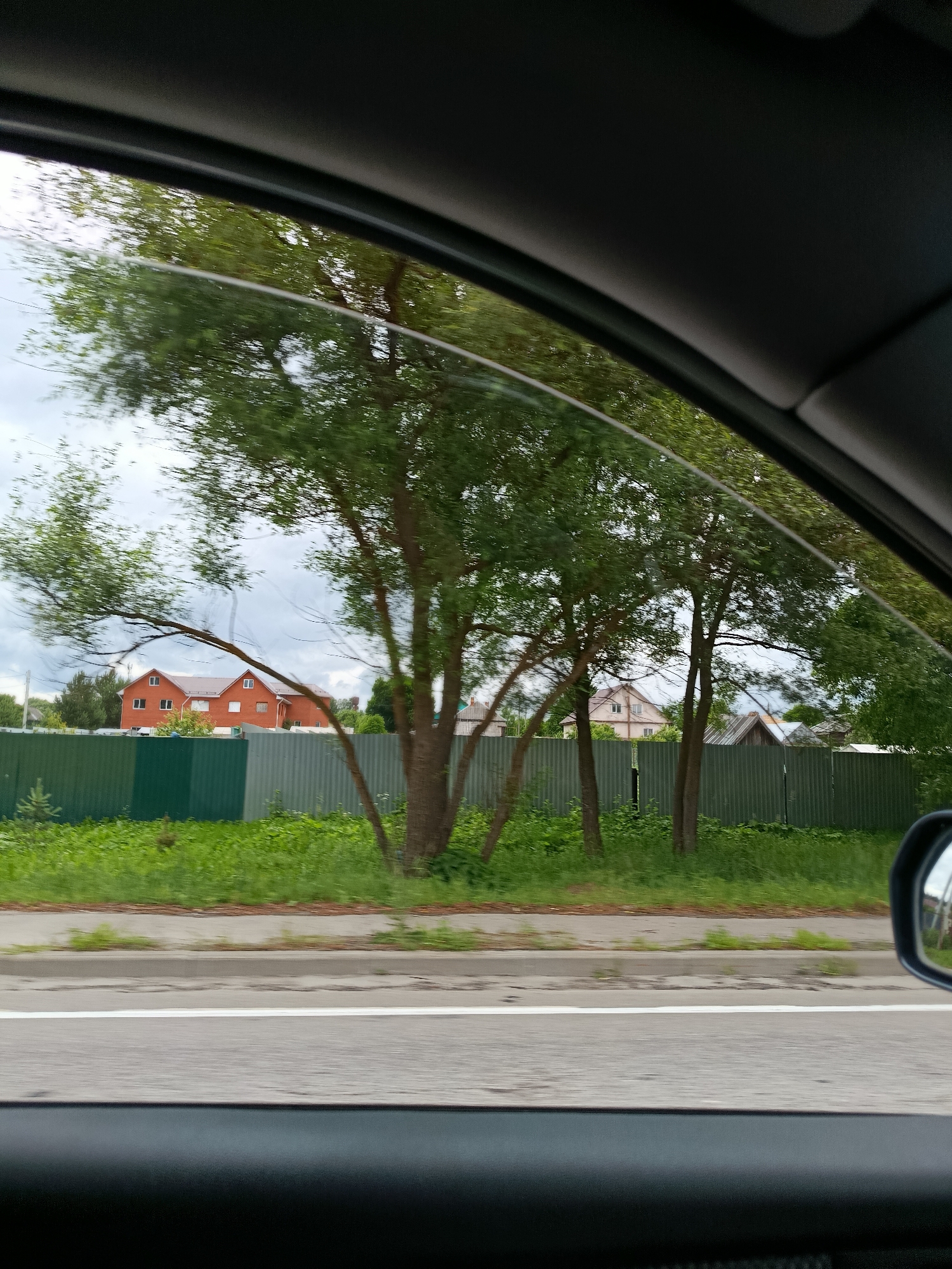 The image is a view from inside a vehicle looking out through the passenger window. The scene outside depicts a suburban or rural setting with a road in the foreground, a green corrugated metal fence, and several residential buildings behind the fence. Trees with green foliage are scattered throughout the scene, some obscuring parts of the buildings. The sky is overcast and cloudy. The location is identified as Savinskaya, Russia.