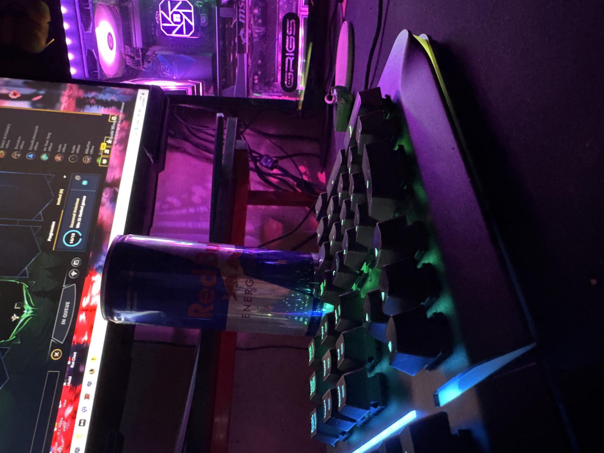 The image depicts a close-up, low-angle view of a gaming setup in what appears to be a residential setting, possibly Varna, Bulgaria, given the hints. In the foreground, a black keyboard with illuminated green and blue keys is prominently displayed, with the keys showing legends like "Insert," "Delete," and "Home." To the right of the keyboard, the edge of a dark mousepad is visible.
Behind the keyboard, a tall, blue and red can of Red Bull Energy Drink is positioned vertically, its metallic surface reflecting the ambient neon lighting. Further in the background, a computer monitor displays a game interface, with text such as "IN QUEUE," "Progression," and player names visible. The monitor screen and the surrounding environment are bathed in vibrant purple and pink neon lighting, characteristic of a gaming setup.
To the right of the monitor, a gaming computer tower is visible, also illuminated with purple LED lighting, featuring a fan with a similar lighting effect. The overall environment is dimly lit, emphasizing the glow of the computer components and keyboard. The composition suggests a focus on the gaming experience, with the energy drink present as a common accompaniment. The image's framing and lighting create a dynamic and immersive atmosphere. There are no people visible in the image.