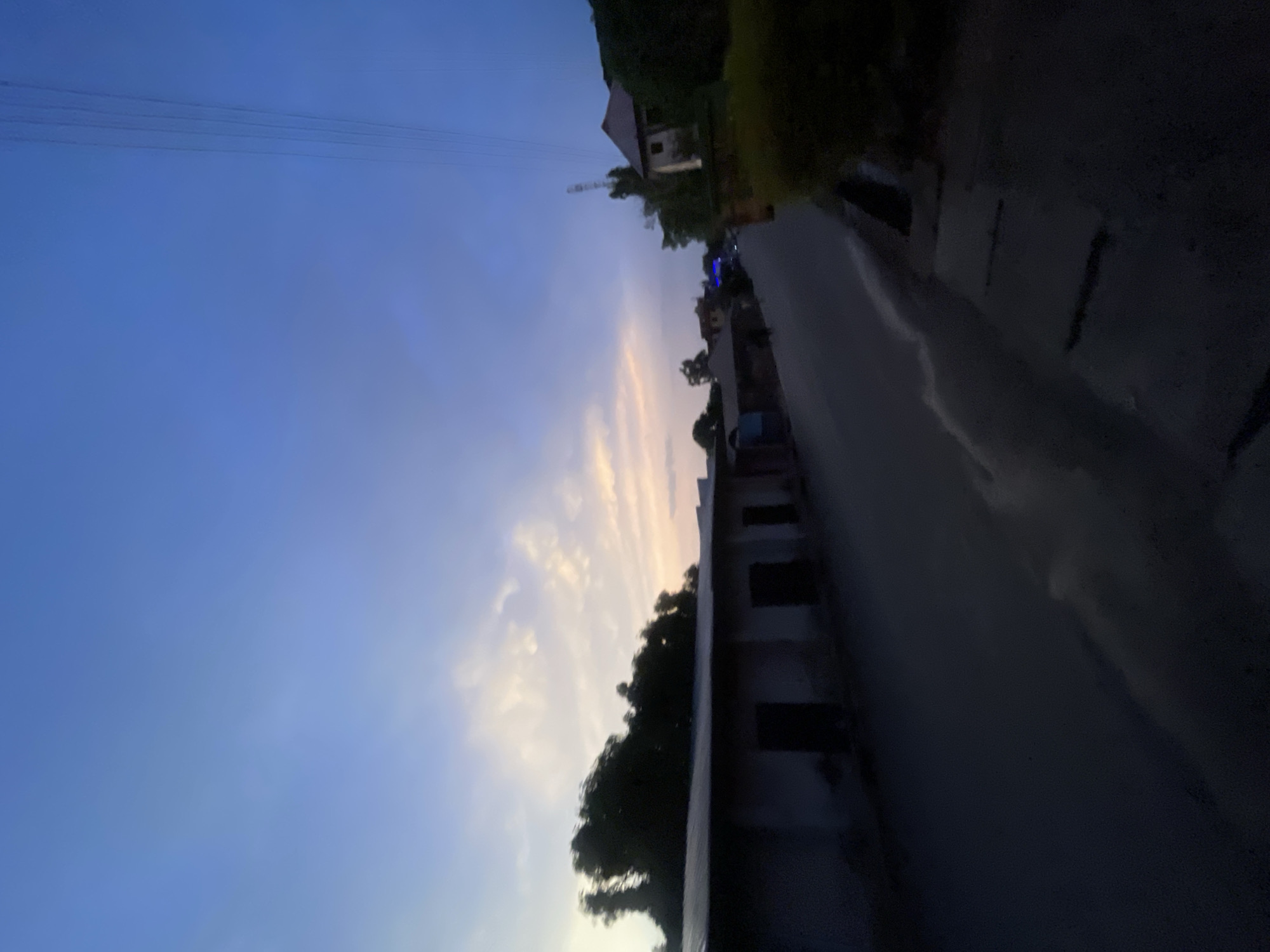 The image depicts an evening scene in Jos, Nigeria. A road stretches into the distance, flanked by buildings on one side and trees on the other. The sky is a gradient of blue and pale orange, possibly indicating sunset or sunrise. Power lines are visible against the sky. The foreground is dimly lit, obscuring details of the road surface and nearby structures.