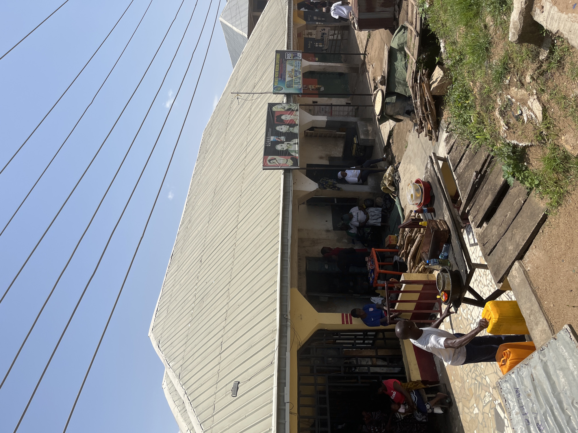 The image depicts a street scene in Gwong, Nigeria, featuring a building with a corrugated metal roof, a common architectural style in the region. Several people are visible in and around the building, which appears to house various businesses or shops.
In the foreground, a man in a white t-shirt and dark trousers is attending to two large yellow jerrycans. He is holding onto one of them, suggesting he might be filling or moving it. Nearby, on a wooden table, are various containers and items, including a red bucket with yellow contents, bottles of water, and what appear to be cooking utensils. Piles of firewood are also stacked in the area, indicating potential cooking or heating activities.
Further back, under the building's overhang, several individuals are seated or standing. Some appear to be customers or residents, engaged in conversation or resting. Advertisements and signs are displayed on the building's facade, including one that reads "BONNY VINCENT SUPER MARKET" and another for "MIFIED REST PATENT MEDICINE." The overall environment suggests a bustling, everyday scene in a Nigerian urban or semi-urban setting. The sky is clear and blue, indicative of sunny weather.