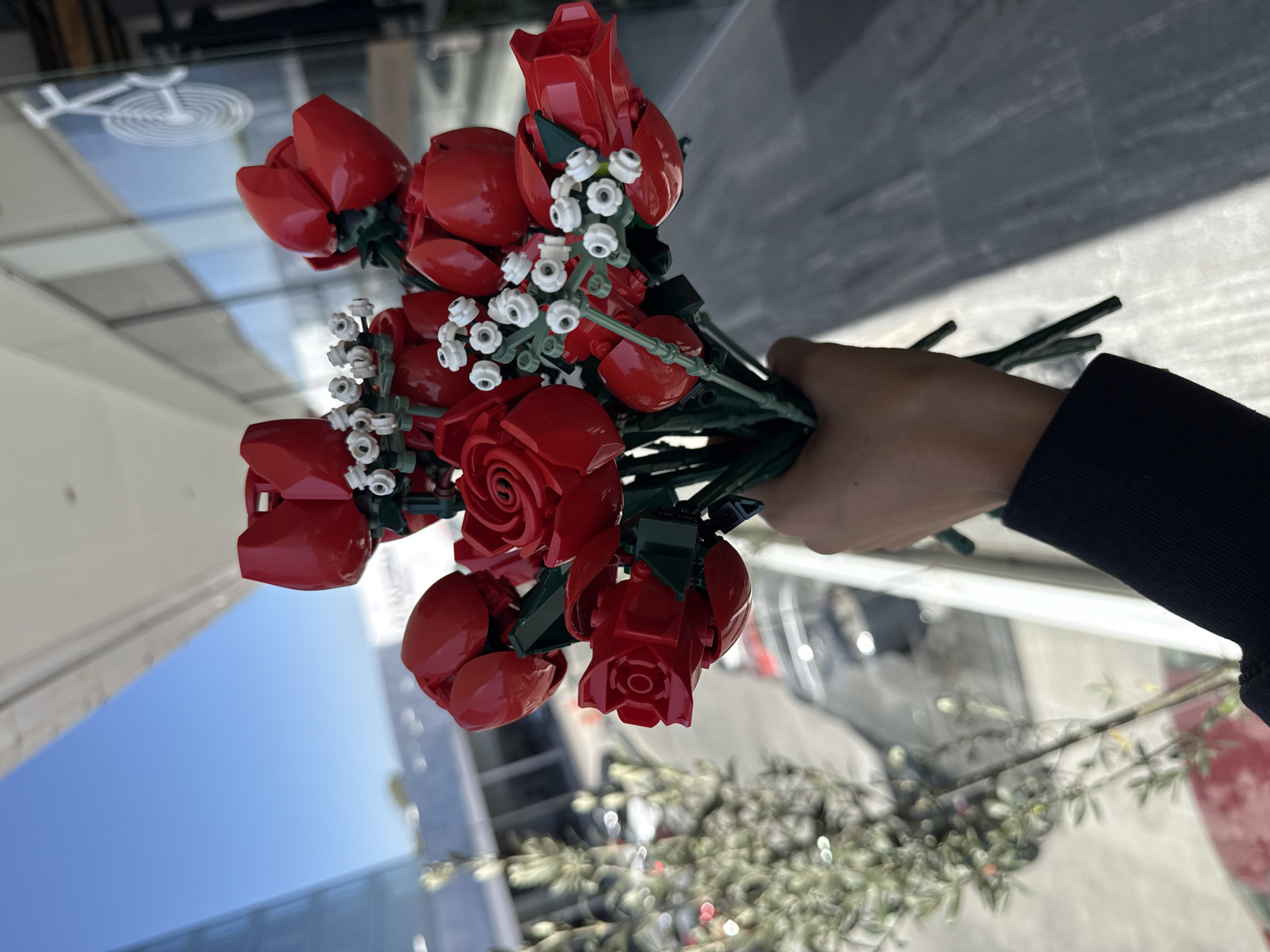 A person's light-skinned hand, wearing a dark long-sleeved garment, holds a bouquet of red roses constructed from LEGO-style bricks. The bouquet features numerous red rose blossoms with green stems and intricate white floral accents resembling baby's breath, also made from building blocks.
The scene is set outdoors in what appears to be an urban or commercial environment, under bright daylight. A clear blue sky is visible in parts of the background, suggesting sunny weather. The background is dominated by large, reflective glass surfaces that show distorted reflections of modern buildings and other elements of the surrounding cityscape. Notably, a white bicycle icon, possibly a decal or stencil, is reflected prominently on a glass surface in the upper left portion of the image. Further details in the background, such as blurred vehicles and some greenery at the bottom, are out of focus, contributing to the sense of a busy public space. No discernible text is visible in the image.