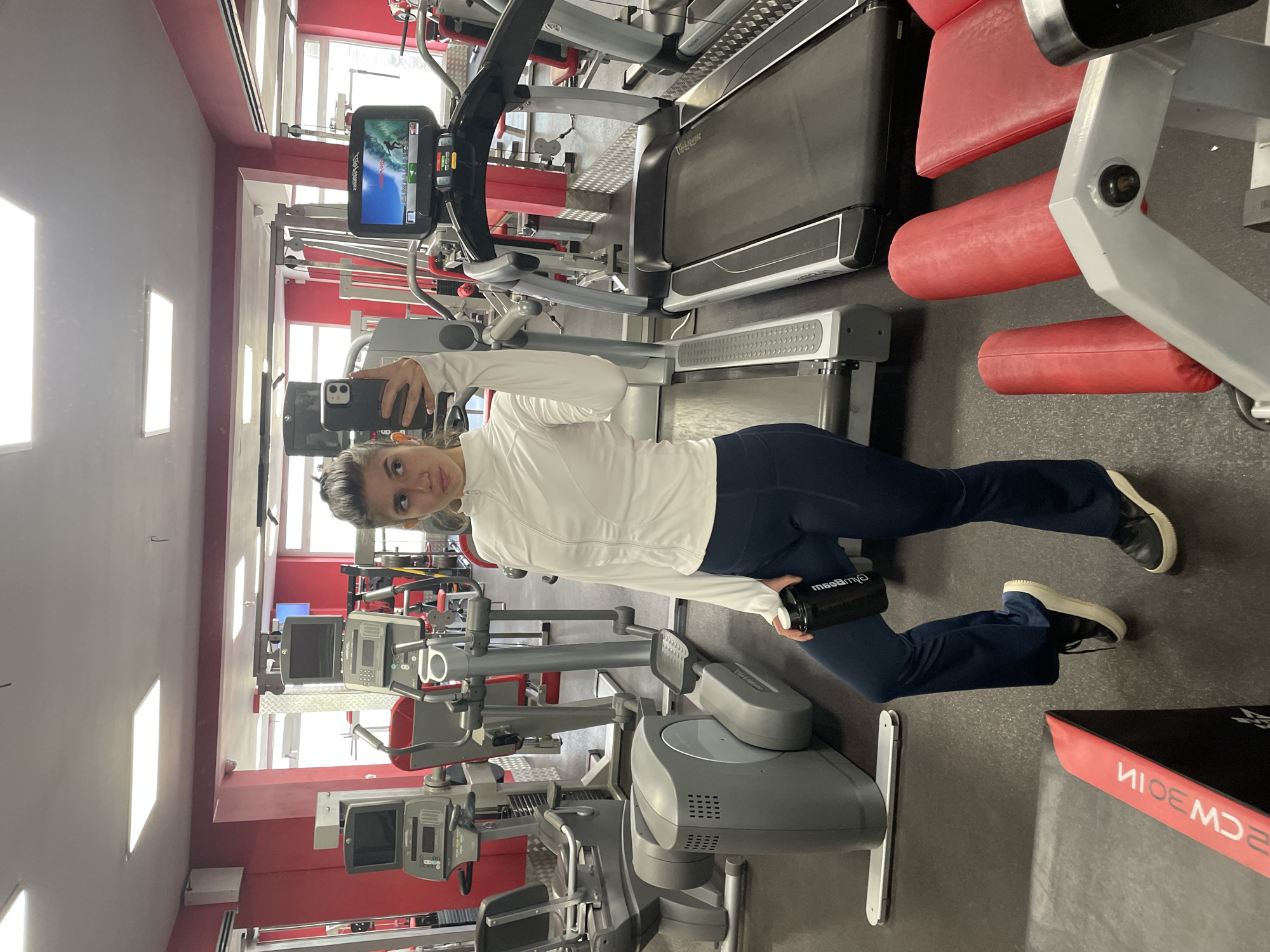 A woman is taking a selfie in a gym. The gym features treadmills and other exercise equipment. The environment appears to be indoors. It is not possible to independently confirm the location as Sofia, Bulgaria from the image alone.