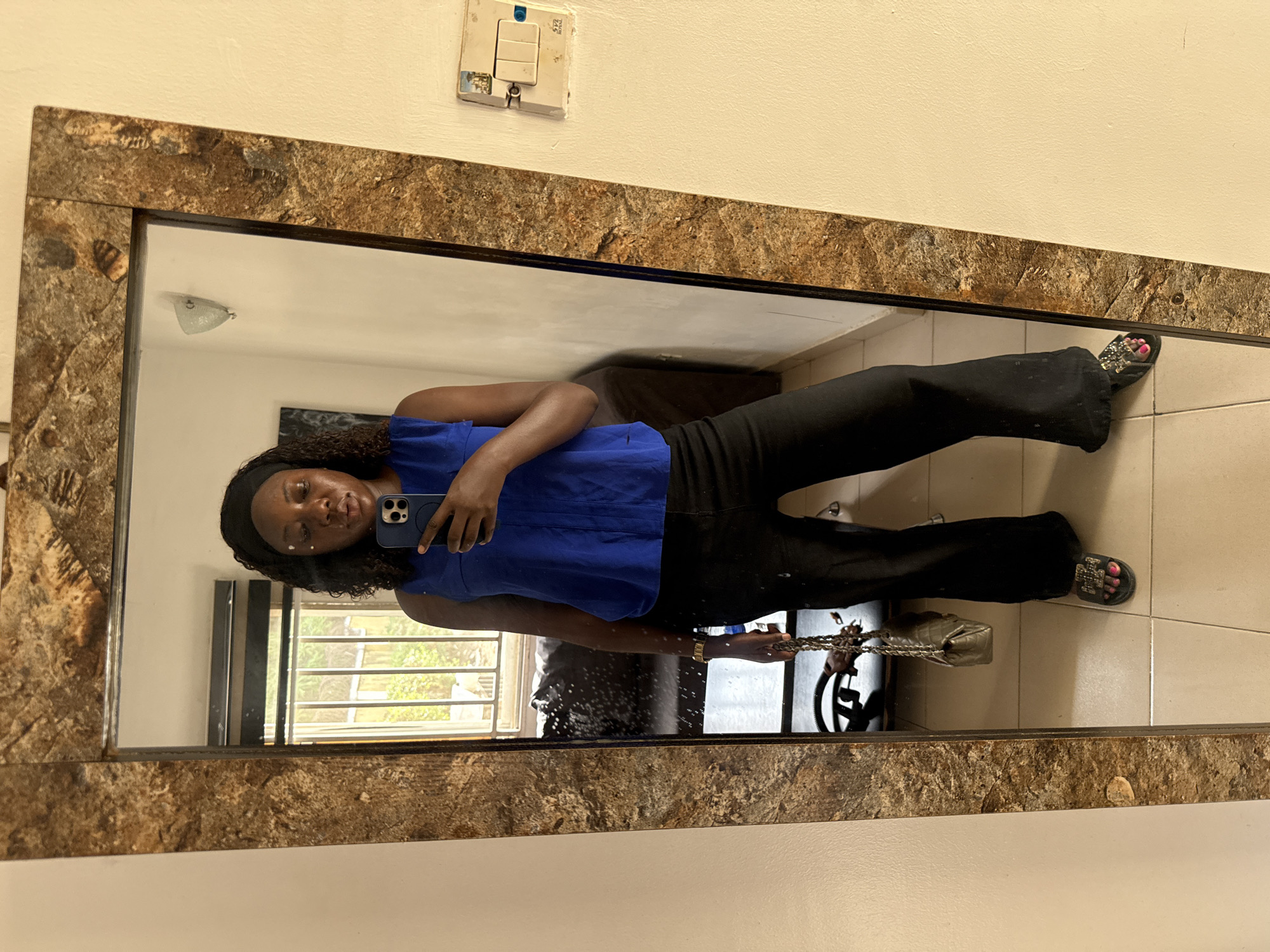 A full-body mirror selfie captures a woman standing upright, holding a smartphone to take her picture. She is positioned centrally within the mirror's reflection.
**Subjects:** The primary subject is a woman with dark, curly hair, pulled back by a black headband. She has a calm, neutral expression, and small white dots of makeup are visible under her eyes.
**Clothing and Accessories:** She wears a vibrant blue sleeveless top paired with black, likely flared, trousers. A gold-colored watch adorns her left wrist. In her right hand, she holds a small, metallic silver or gold handbag with a chain strap. Her feet are clad in black sandals featuring sparkling embellishments, and her toenails are painted bright pink.
**Objects:** Prominent objects include the woman's smartphone (appearing to be an iPhone), her handbag, and the large rectangular mirror. The mirror features a thick frame with a textured, mottled brown and gold pattern, resembling stone or marble. Above the mirror on the cream-colored wall, an older-style white light switch and power outlet unit is mounted.
**Scene and Setting:** The photograph is taken indoors, in what appears to be a room with cream-colored walls and a light-tiled floor. The mirror's reflection shows a window with dark horizontal and vertical bars, through which green foliage and bright daylight are visible, suggesting an outdoor view. A ceiling light fixture is also reflected in the upper portion of the mirror.
**Activity:** The woman is actively taking a mirror selfie, looking at her phone.
**Notable Details:** The time of day appears to be daytime, indicated by the natural light filtering through the window. The light switch on the wall has partially visible text that reads "LG" and "SCALA". The overall scene suggests a casual, everyday moment in a home or similar indoor setting in Abuja, Nigeria.