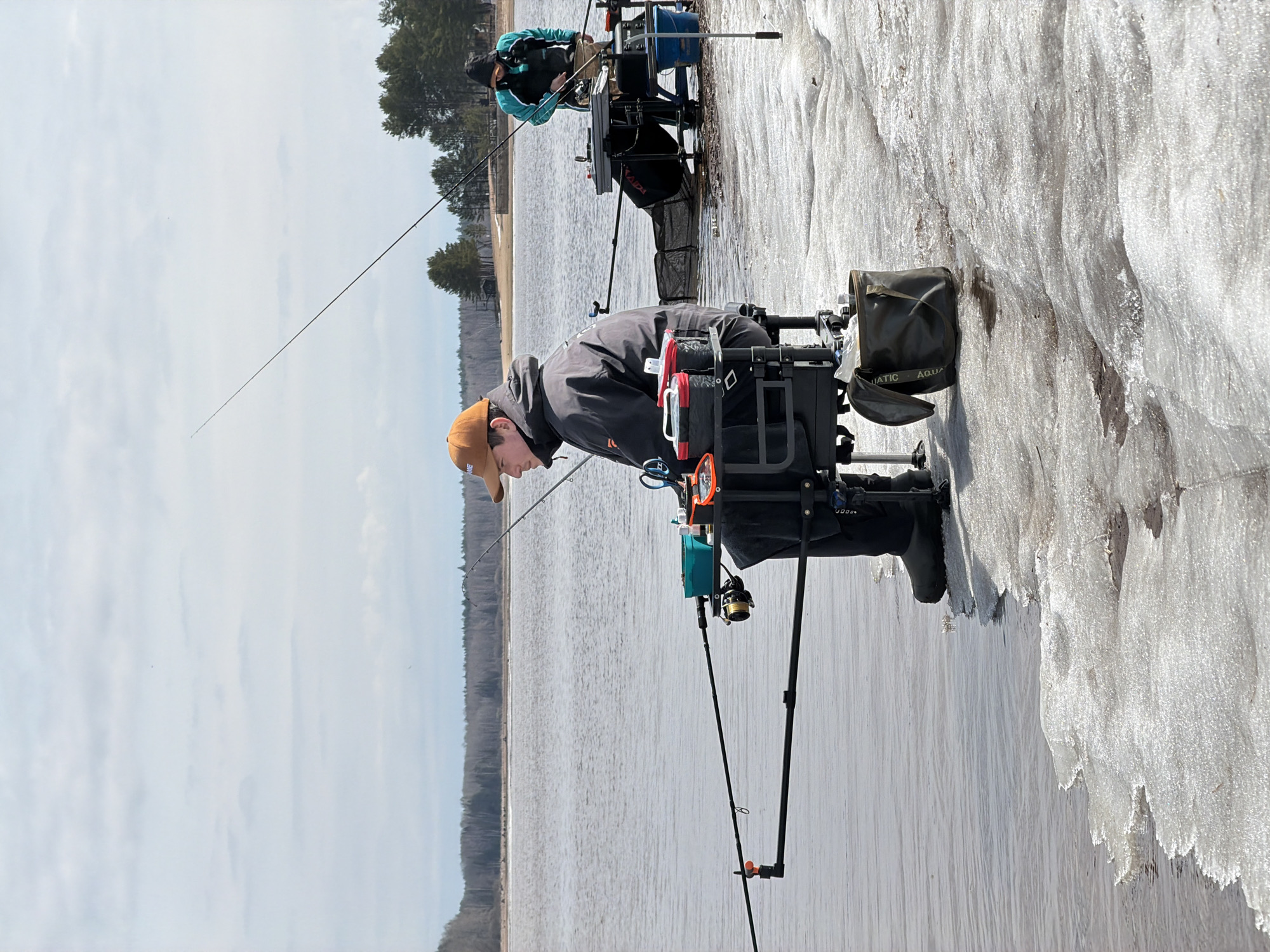 Two people are ice fishing. The location is most likely in the vicinity of Chaykovsky, Russia, based on the presence of pine trees and a large body of water. The foreground shows a man in a dark jacket and tan hat fishing with specialized equipment, including a fishing rod stand attached to his seat. Another individual is fishing further in the background. The scene is set on a partially frozen body of water, with ice formations along the edge. The sky is overcast.