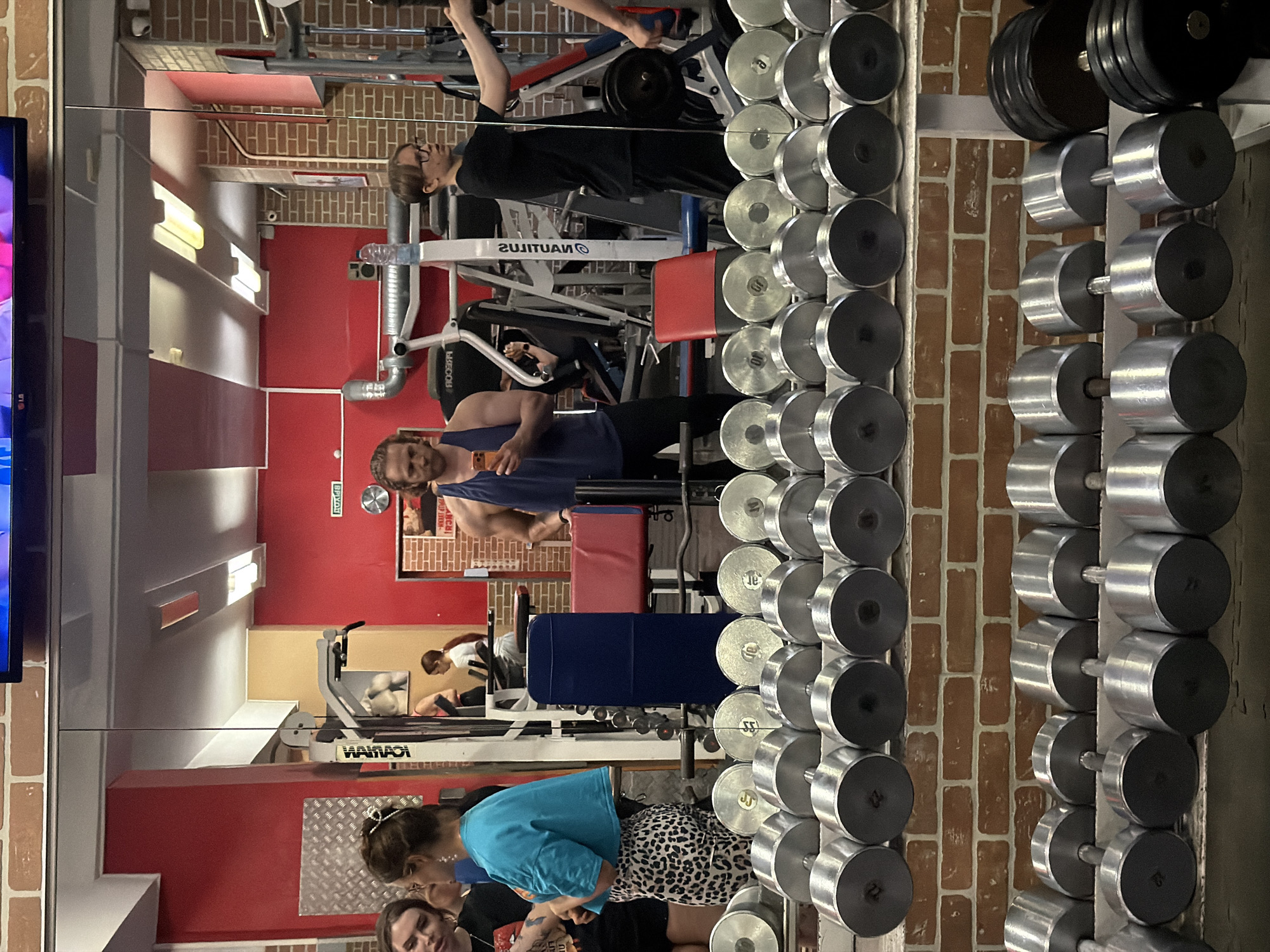 The image is of a gym, with a mirrored wall showing reflections of the space and people within it. The location is Saint Petersburg, Russia, as indicated by the decor and architecture.
Several individuals are present. One man, reflected in the mirror, wears a blue tank top and appears to be holding a phone. Another man in black is using a Nautilus gym machine. Another group of people is closer to the camera, including a woman in a blue shirt and leopard print pants. Other people are vaguely visible.
The gym contains various fitness machines and equipment, including a weight rack with a variety of dumbbells, as well as Nautilus machines. The walls are partially brick and partially painted red and tan. There is a television on the far-left side of the image.