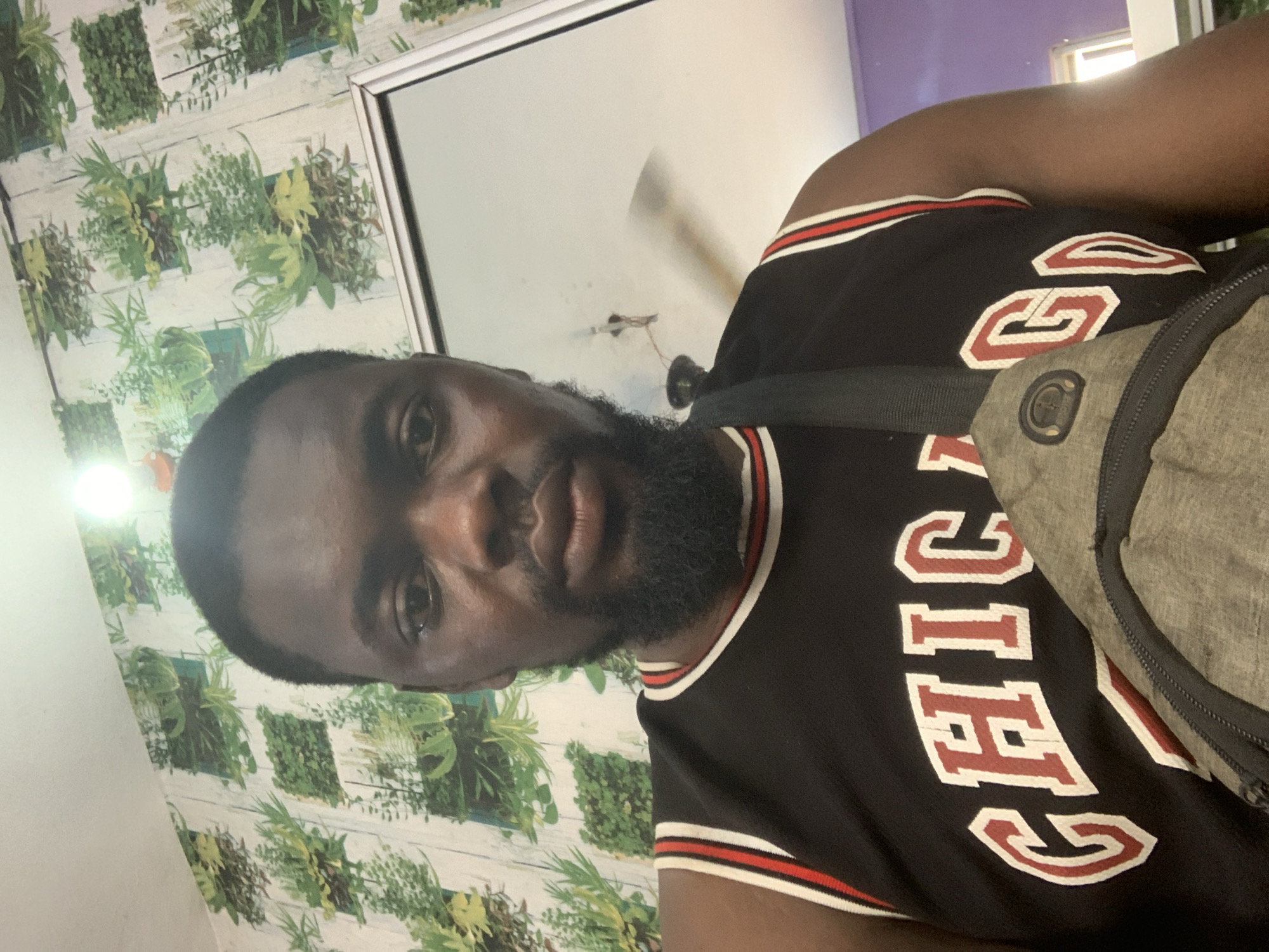The image is a selfie of a Black man wearing a black Chicago Bulls basketball jersey with red and white trim. He has a beard and is wearing a gray crossbody bag. The man is standing in front of a wall with a botanical print wallpaper. A ceiling fan is visible in the background, suggesting an indoor environment. The lighting appears to be from an overhead lamp. The scene is not identifiable as being in Shagamu, Nigeria.