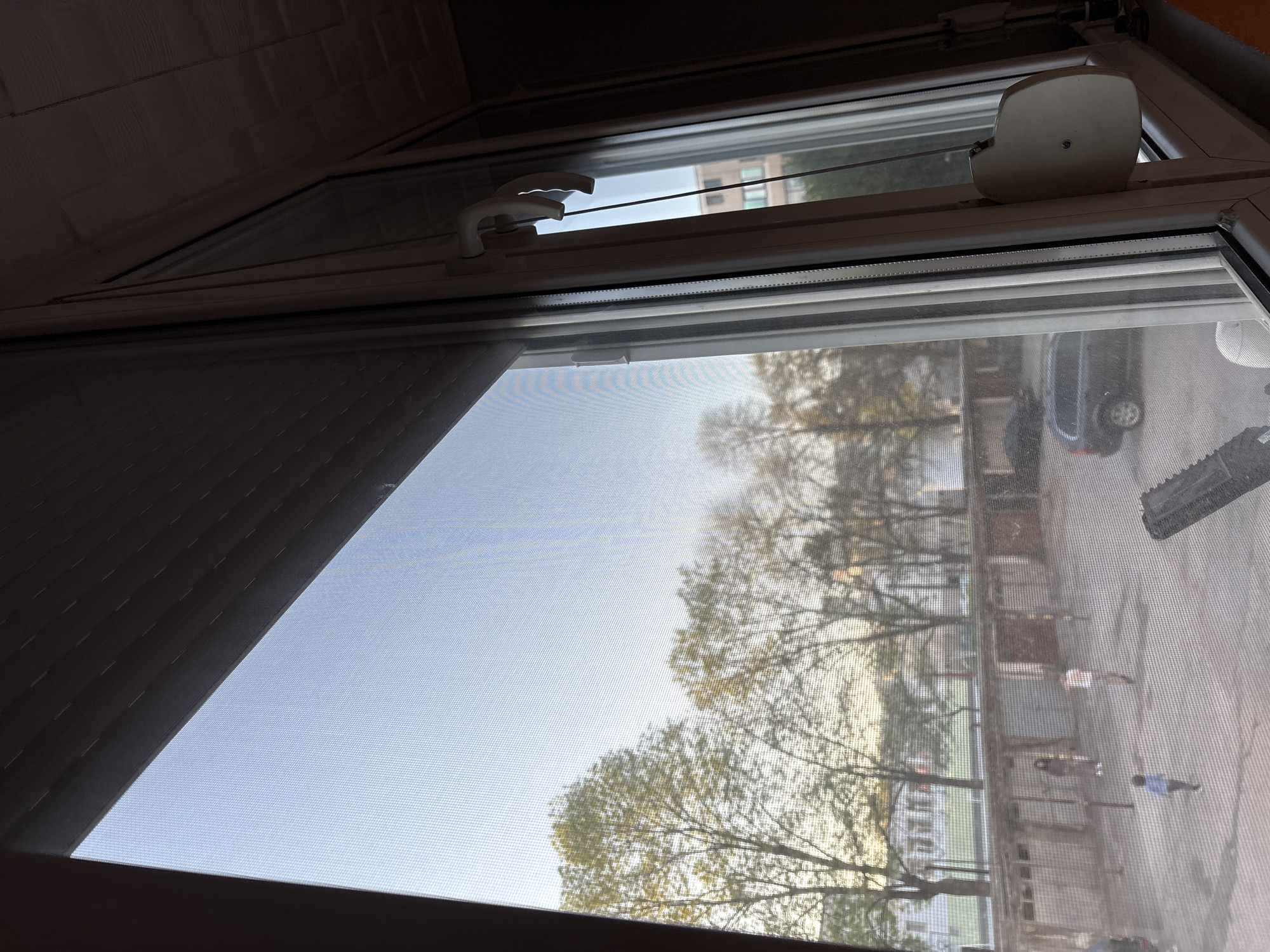 View from an open window in Panagyurishte, Bulgaria. The window has a screen. Outside, trees are visible. Several people are walking along a sidewalk. A parked car is visible.