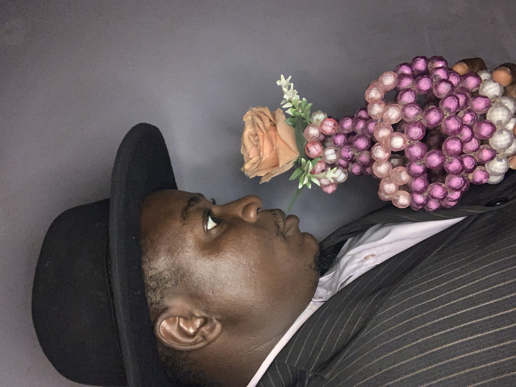 A dark-skinned adult individual, observed in right profile, wears a black bowler hat, a dark pinstriped suit jacket, and a light-colored collared shirt. He possesses a trimmed mustache and beard. His gaze is directed downwards towards an object held in his hands, which consists of a peach-colored rose, small white florets, and a substantial cluster of faceted spherical beads in shades of pink, purple, and white. The individual appears to be smelling or closely examining the rose. The background is a plain, uniform grey wall. There are no visual cues within the image to indicate its location in Akwanga, Nigeria, or any other specific geographical setting.