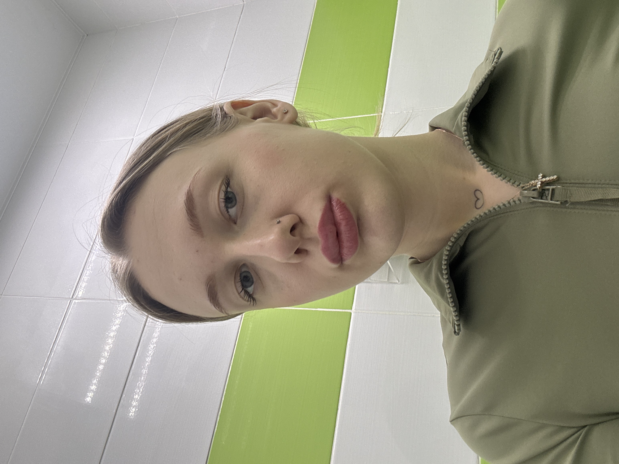 A young adult female is depicted in a close-up selfie, facing the camera with a direct gaze and pursed, pink-lipsticked lips. She has light hair pulled back from her face, light-colored eyes, and a small stud piercing in her left nostril. A small, simple heart outline tattoo is visible on her left upper chest/neck area, accompanied by a thin gold-colored chain necklace with a small cross pendant. She is wearing an olive green or khaki-colored quarter-zip collared top. The background consists of a glossy tiled wall featuring horizontal bands of white and bright green rectangular tiles. This scene is located in Rostov-on-Don, Russia.
