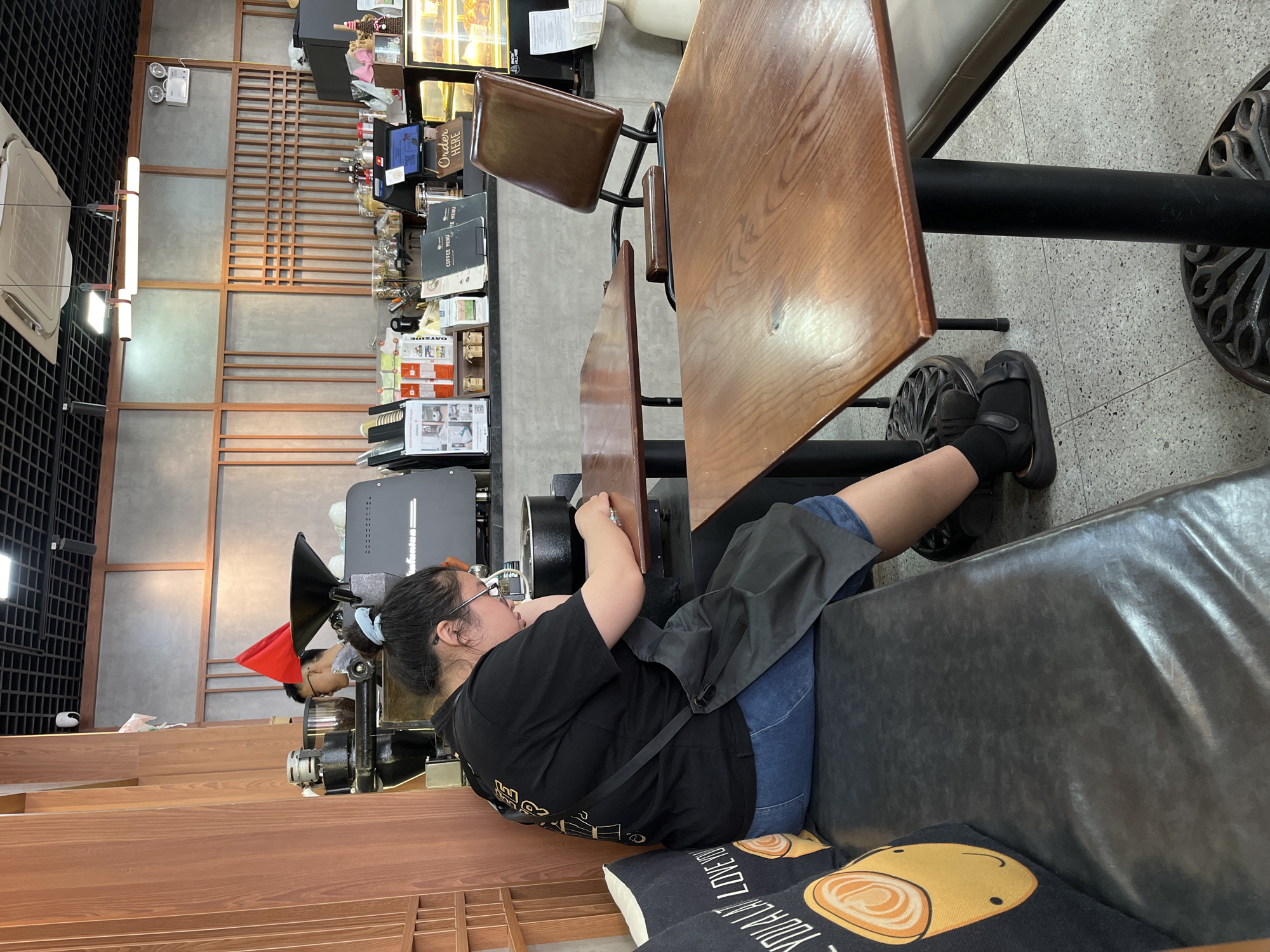 A woman is sitting on a black bench in what appears to be a café or restaurant. She has dark hair pulled back, is wearing glasses, a black t-shirt, blue denim shorts, and black sandals with black socks. She has a dark apron on. The woman is turned towards the left of the image and seems to be looking at and working on something just in front of her.
Next to the bench is a wooden table with two chairs. The floor is light grey and appears to be concrete. Behind the bench and woman is a wooden structure. Further behind is a coffee roaster with a person in a red hat standing next to it, and a wall. To the right of the roaster is a display case with food and menus behind the table.
The photograph appears to have been taken indoors. There is a pillow beneath the woman. The image does not contain definitive information that confirms it is in Đà Nẵng, Vietnam.