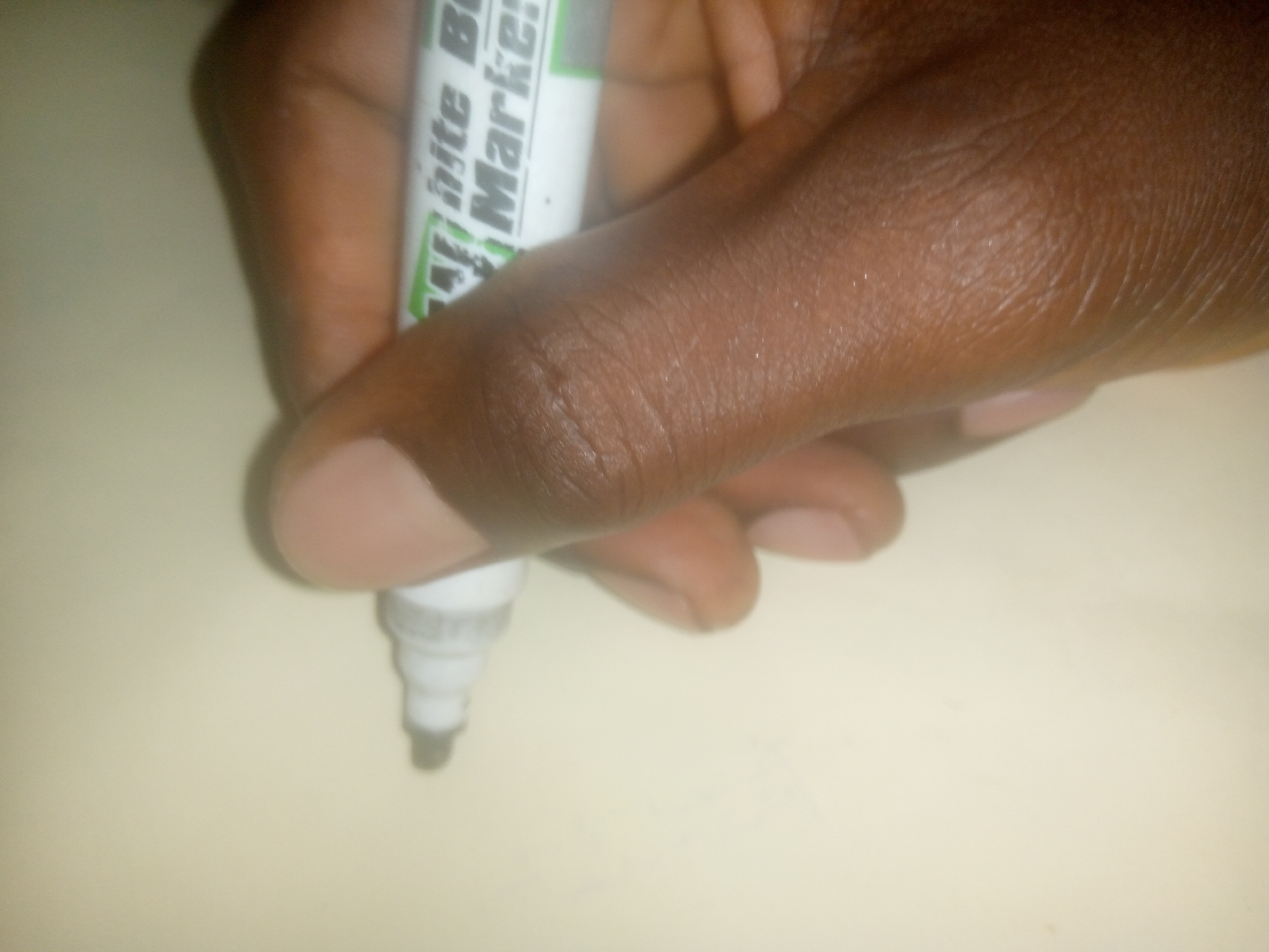 A close-up, angled shot shows a dark-skinned hand holding a white dry-erase marker. The marker has black lettering on a green stripe, which appears to read "White Bo... Marker". The marker tip is pointed downwards and is out of focus. The background is a light-colored, textured surface, possibly a desk or paper. The image is brightly lit, with some specular highlights on the hand and marker. The scene is neutral and does not suggest any specific location within a city.