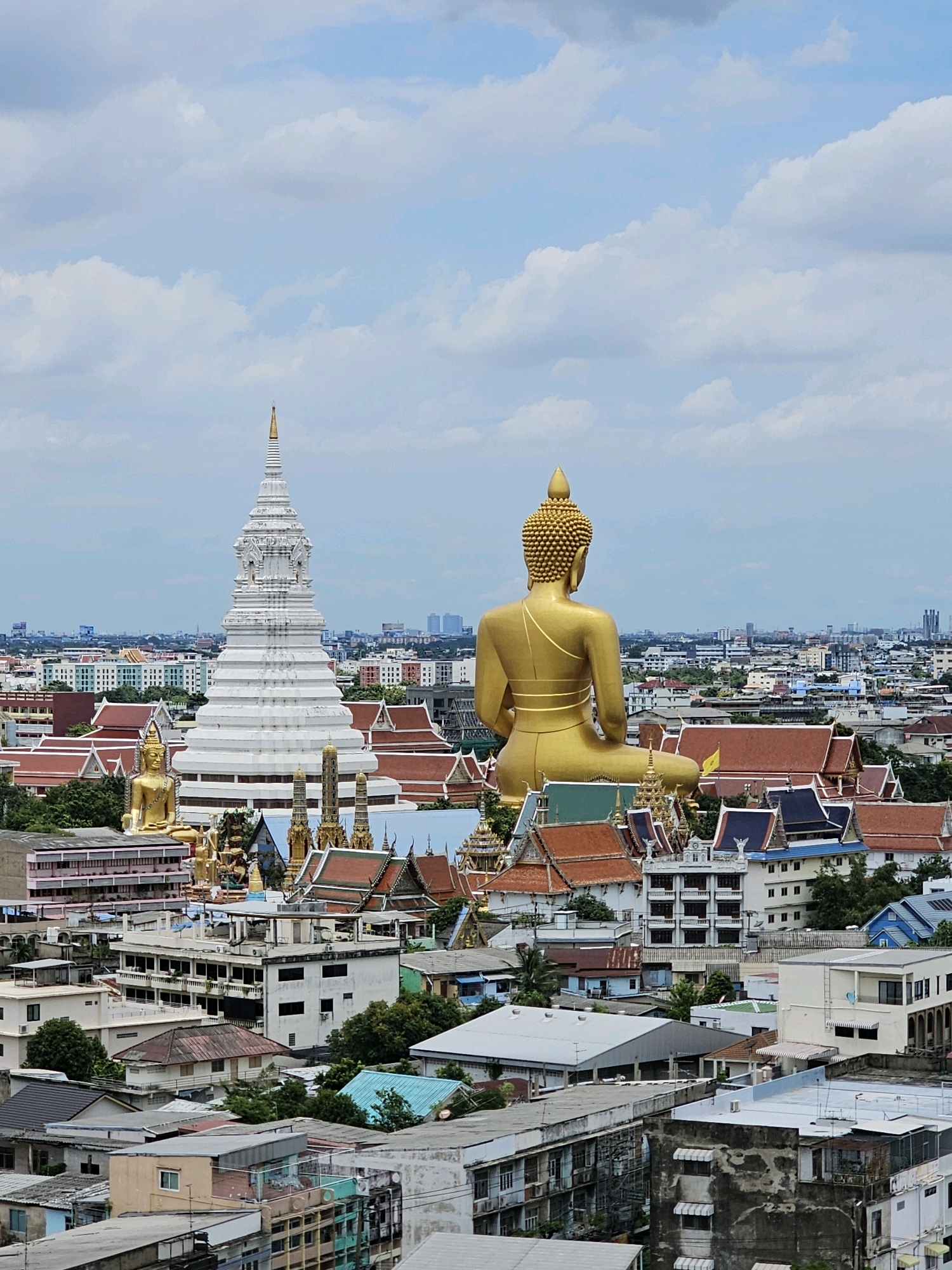 The image depicts a cityscape in Bangkok, Thailand, dominated by a massive golden Buddha statue seated in the foreground, facing away from the camera. Behind the golden Buddha, a large white tiered stupa stands prominently, adorned with intricate carvings and a golden spire. The surrounding area is a dense urban landscape with a mix of traditional Thai temple architecture, characterized by orange-tiled roofs and ornate structures, interspersed with modern multi-story buildings and houses. The sky above is a pale blue with scattered white clouds. To the left of the main golden Buddha, a smaller golden Buddha statue is visible, also seated. The overall scene captures a blend of religious landmarks and urban development in Bangkok.