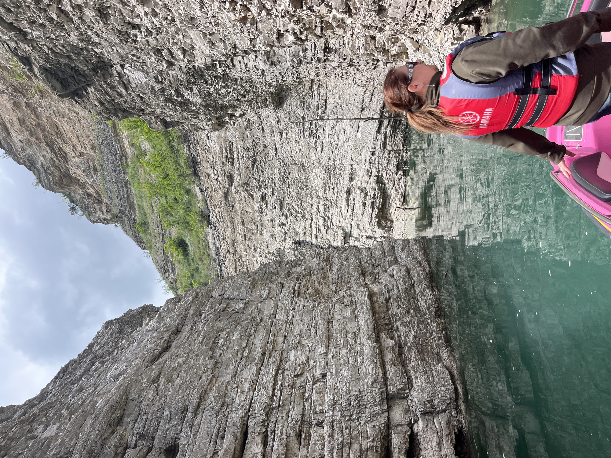 A woman wearing a red and blue life jacket sits in a pink kayak on a body of water. Towering rock formations line the water's edge. The sky is overcast. The brand "Yamaha" is visible on the life jacket. A city of Druzhba, Russia, location cannot be confirmed.