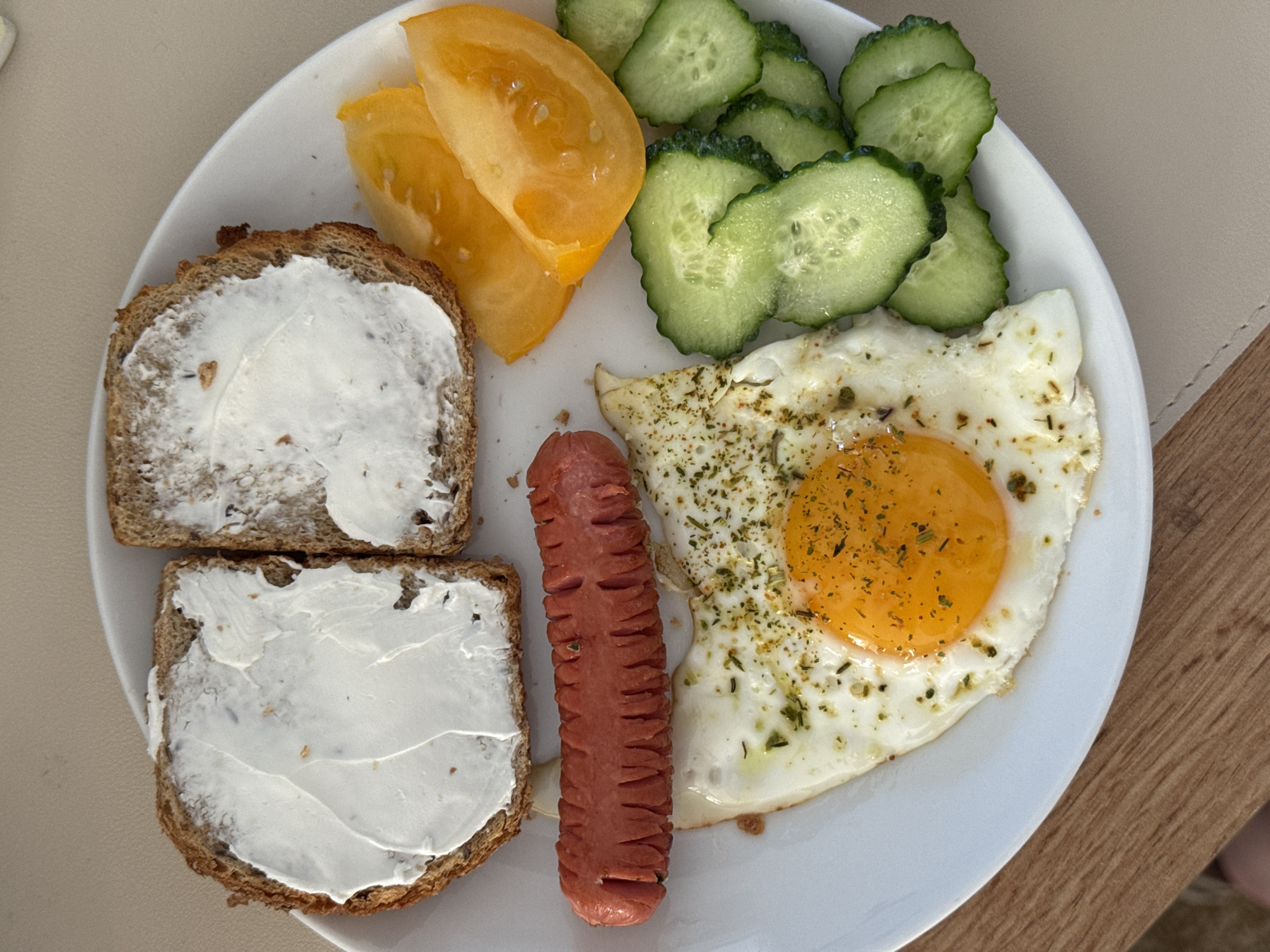 A close-up overhead shot shows a white plate of breakfast food on a beige placemat and a wooden surface. The plate contains two slices of whole wheat bread spread with a white substance, possibly cream cheese. Next to the bread is a grilled sausage with cuts that create a zig-zag pattern. A fried egg with a bright orange yolk, seasoned with green herbs, is positioned below the sausage. To the right of the egg are sliced cucumbers and wedge-shaped slices of yellow tomato. The background is out of focus, but appears to be an indoor setting.