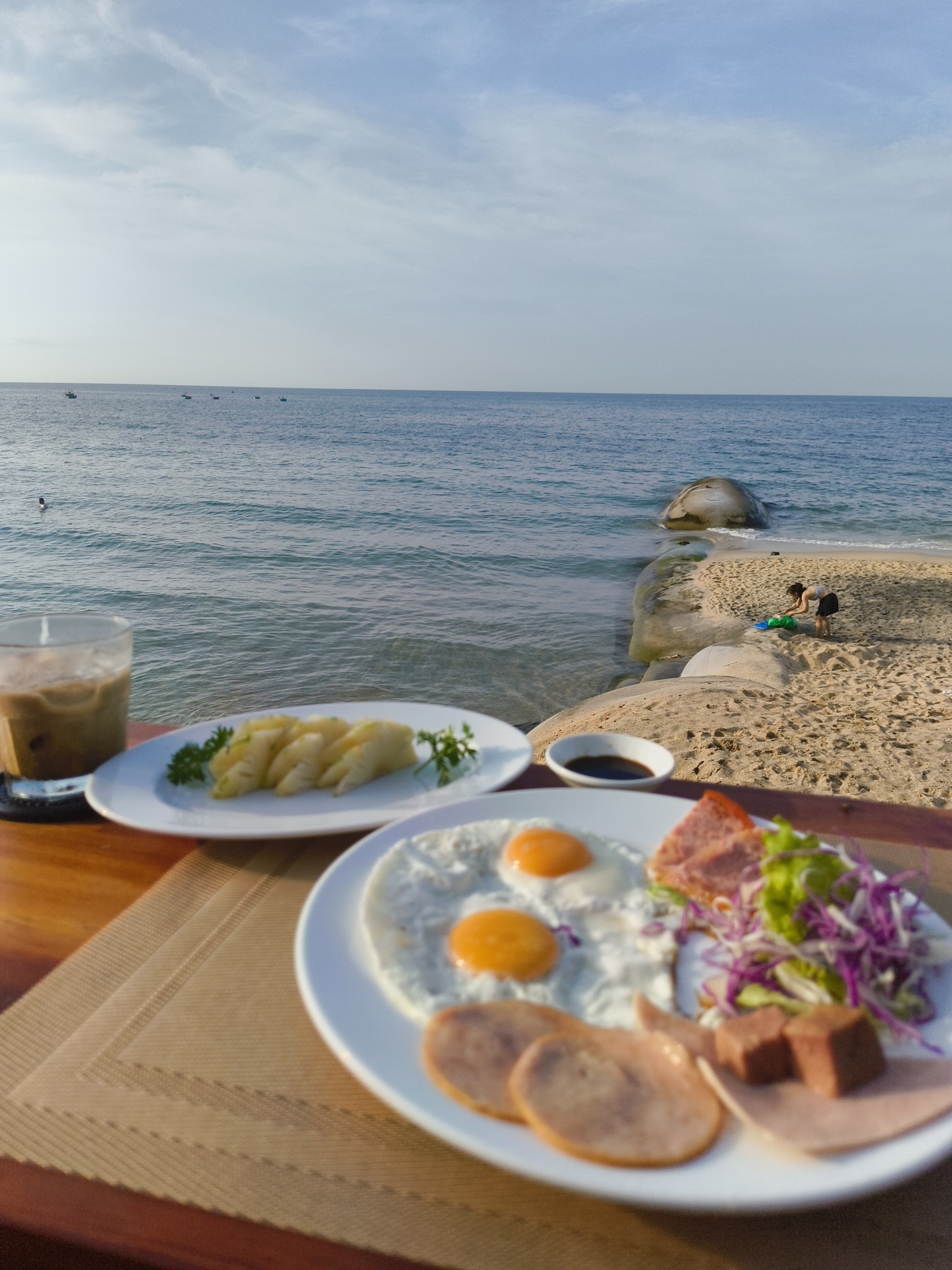 In Phan Thiết, Vietnam, a breakfast spread is laid out on a wooden table beside a tranquil beach. The ocean, with calm, blue water and a few distant boats, stretches to the horizon under a pale, cloudy sky. On the sand, a person is bending over, interacting with a green object near large, smooth rocks. In the foreground, a glass of iced coffee sits next to a plate of sliced pineapple garnished with parsley. Another plate displays a breakfast of two fried eggs, sliced ham, and a salad of lettuce and red cabbage. A small dish of dark sauce is also present. The scene captures a peaceful morning by the sea, with the vibrant food contrasting against the serene natural environment.