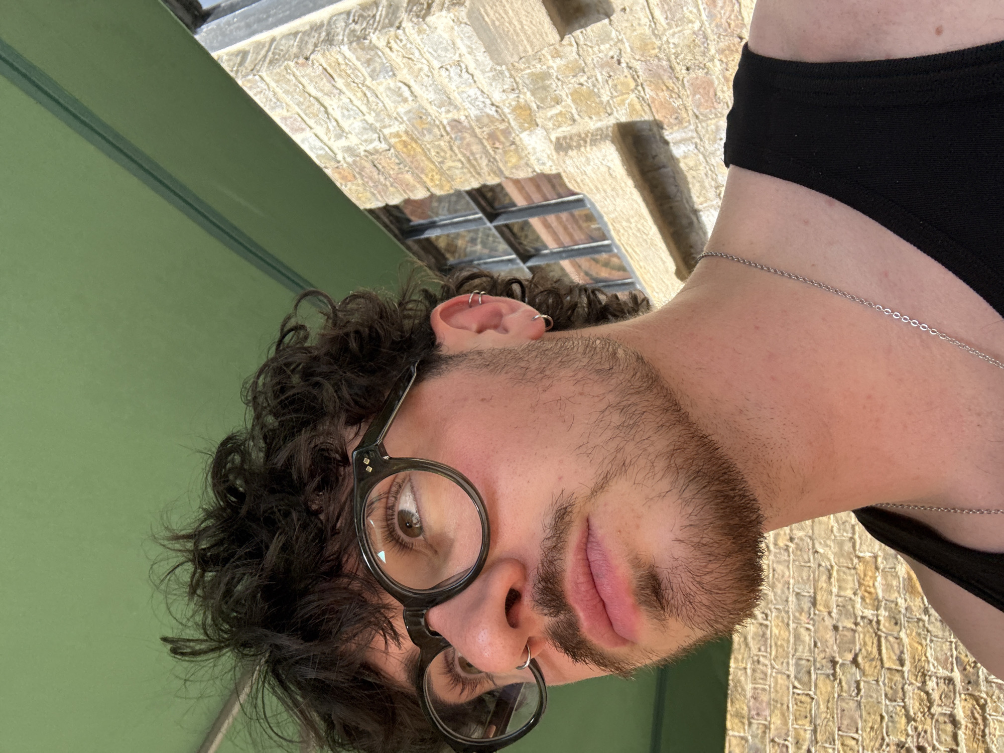 A low-angle close-up portrait shows a man with curly brown hair, wearing round, black-rimmed glasses. He has a light beard and multiple piercings in his ear and nose. A thin silver chain is visible around his neck. He is wearing a black garment. The background includes a green surface and a brick structure with a metal grate. The location is Unknown, Unknown.