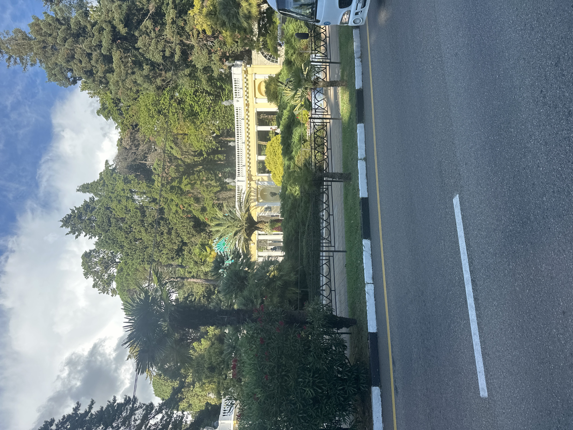 The image shows a view of a yellow building with a white colonnade and decorative elements, set against a backdrop of lush green trees. In front of the building, there is a landscaped area with various plants, including palm trees and bushes, enclosed by a black metal fence. A white vehicle is partially visible on the right side of the frame, suggesting it is parked near the road. The foreground displays a grey asphalt road with white and yellow markings, indicating it is a street or highway. The sky above is partly cloudy with patches of blue. The architecture and vegetation suggest a location in a warm climate, possibly a park or a resort area. Based on the style of the building and the surrounding flora, the location is likely in the city of Sochi, Russia, known for its subtropical climate and numerous parks and recreational facilities.