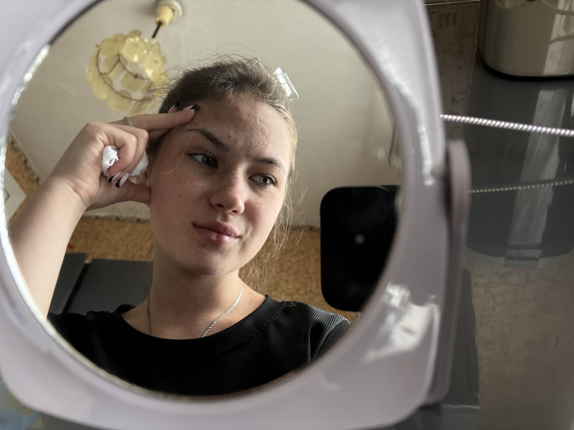 A young Caucasian female is reflected in a mirror, indoors. She has fair skin with freckles and some acne visible on her forehead and cheeks. Her dark blonde hair is pulled back, with a clear plastic hair clip visible at the top of her head. She is wearing a black ribbed t-shirt and a thin silver chain necklace. Her right hand is raised to her forehead, and she is holding a small piece of white tissue in her fingers. Her fingernails are painted black and white. The background reflected in the mirror shows a wall with cork tiling, a light fixture on the ceiling, and other objects that are out of focus. The mirror itself appears to be a portable vanity mirror with a light purple frame, which is partially visible in the foreground of the image. The overall impression is that of a personal moment of self-examination or skincare routine.