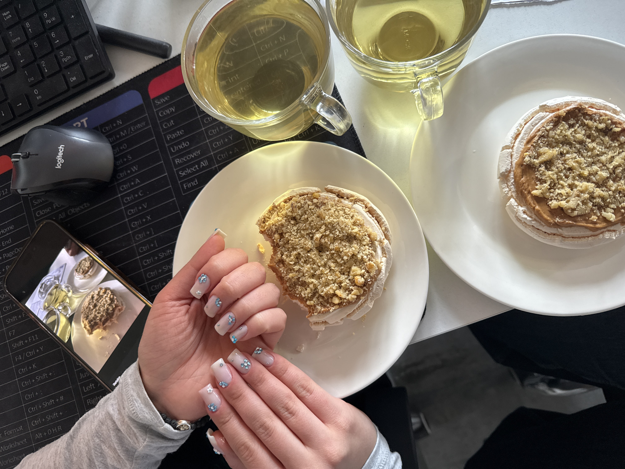 The image shows a desk with a black keyboard and a Logitech mouse on a black mousepad. There are two glass cups of green tea and two plates, each with a meringue-based dessert topped with a brown filling and chopped nuts. A person is holding a smartphone, which displays a photo of the same scene. The person has manicured nails with a white base and small blue flower decorations. The person is wearing a grey long-sleeved shirt and a bracelet. The location in the city of Yerevan, Armenia is not discernible from the image.