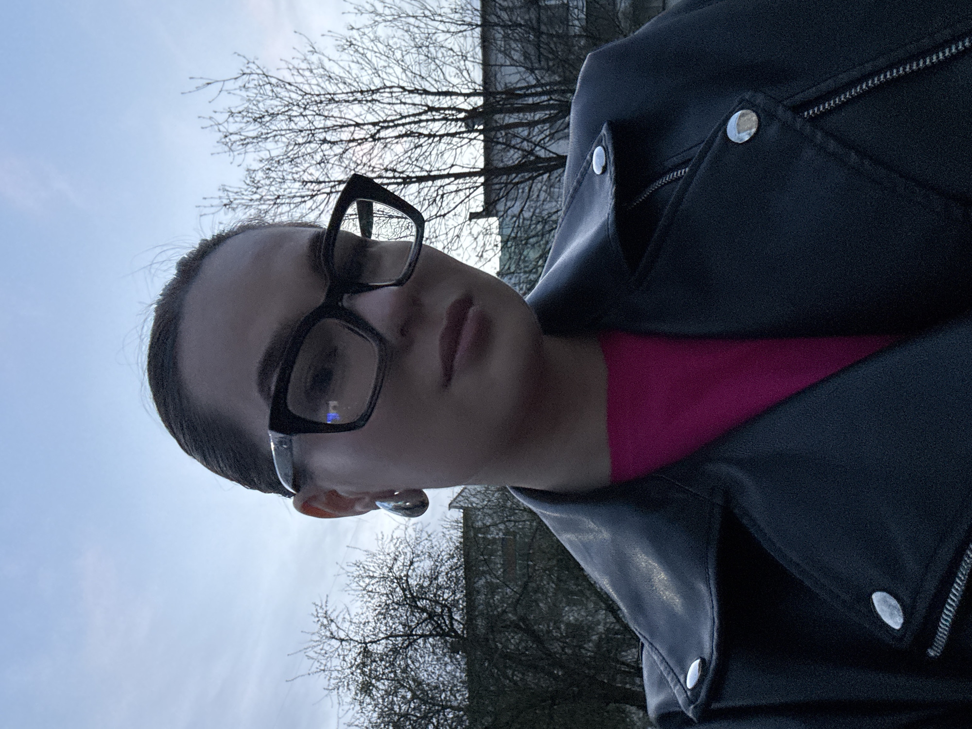 A close-up portrait shows a person, likely female, looking directly at the camera. They wear large, rectangular black-framed glasses that reflect some light, and their hair is pulled back. Underneath a black leather jacket with silver snaps and zippers, a bright magenta or pink garment is visible. The jacket features prominent lapels and a zipped pocket on the chest.
The person's head and upper torso fill a significant portion of the frame, which is oriented vertically, suggesting a selfie or a low-angle shot. The background features a clear, light blue to grey sky, indicating an overcast or cloudy day. Several bare-branched trees, characteristic of autumn or winter, are visible against the sky and behind the person. In the upper background, a dark, indistinct building structure can be seen, suggesting an urban or suburban setting in Chaikovsky, Russia. There is no visible text in the image. The light is diffused, and there are no strong shadows, consistent with an overcast sky.