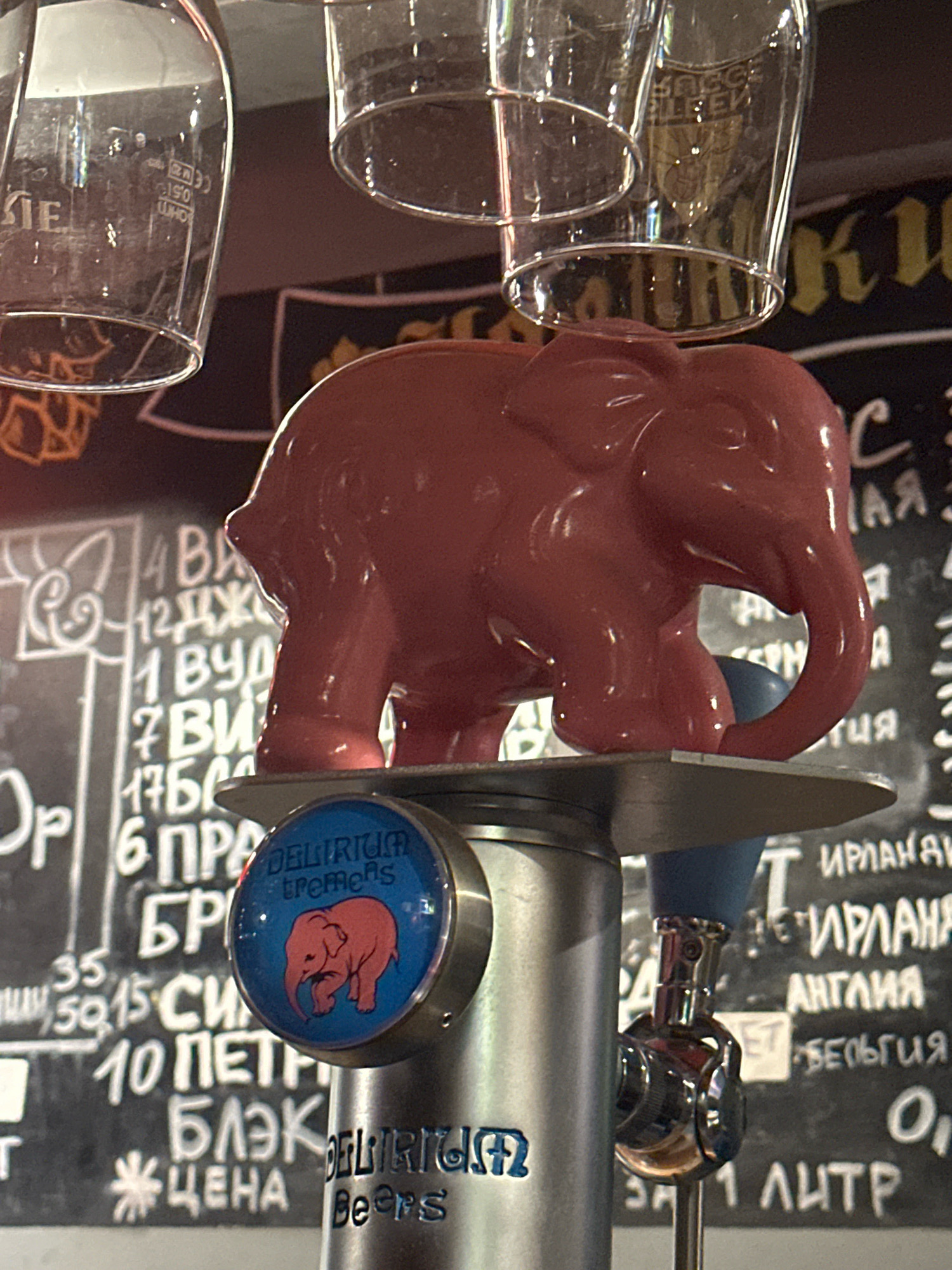A close-up shot captures a pink elephant figurine on top of a beer tap handle. The elephant is positioned centrally in the frame, with its trunk curled downwards. Below the elephant, the blue circular emblem of "Delirium Tremens" beer is visible, featuring a smaller illustration of a pink elephant. The beer tap itself is made of metallic material and appears to be in use. In the background, a chalked menu board displays various beer names and prices in white lettering. The scene is likely in a bar or pub, suggested by the presence of beer taps, glasses hanging in the background, and the menu board.