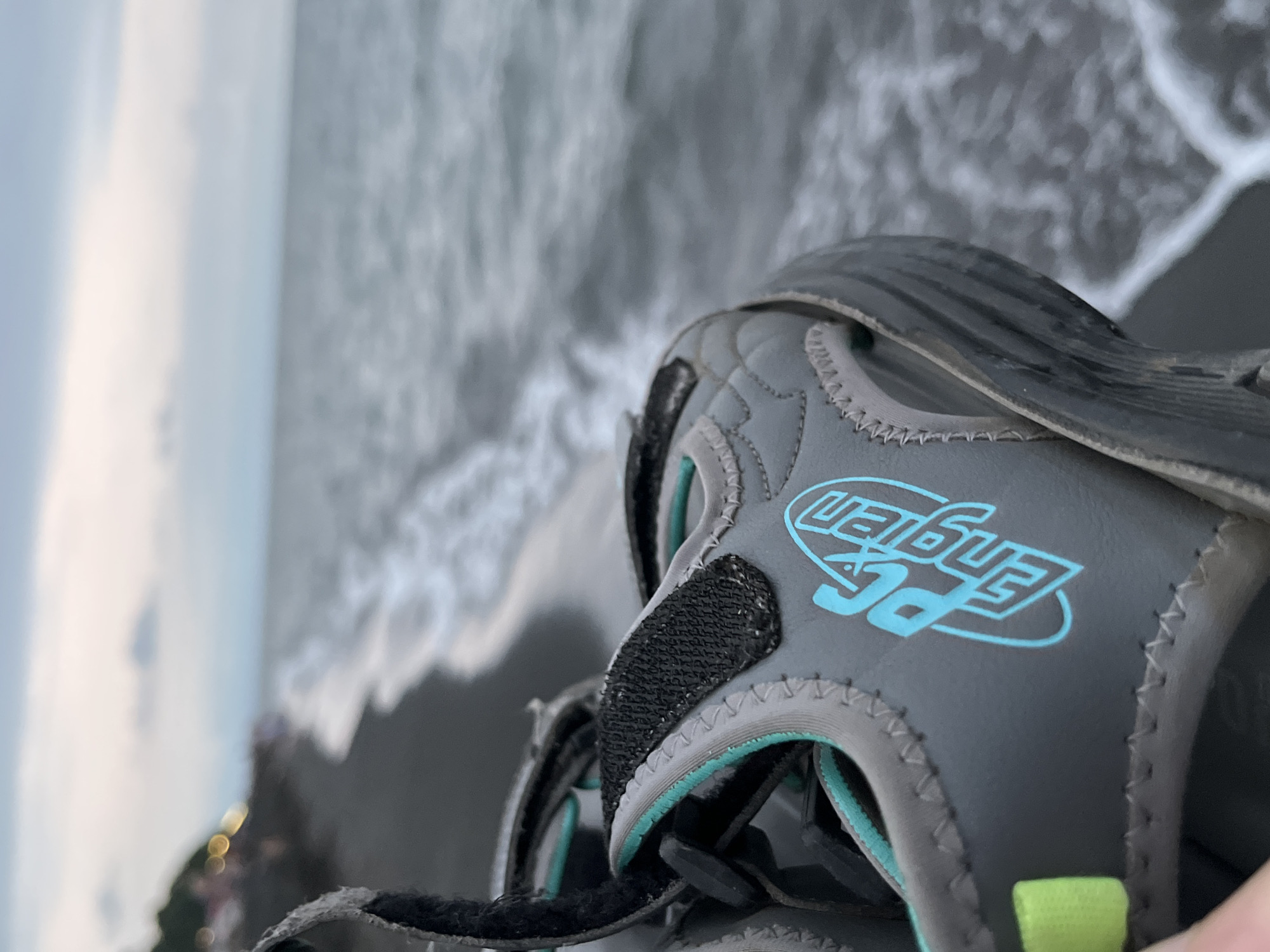 An adult's hand holds a close-up of a grey and teal sandal with a neon green strap, set against the backdrop of the Ureki, Georgia beach. The sandal features the logo "PC Engen" in bright teal lettering. The background shows the dark, wet sand of the beach where the waves are gently rolling in, creating white foam. In the distance, a hazy view of buildings and trees can be seen along the shoreline under a cloudy sky.