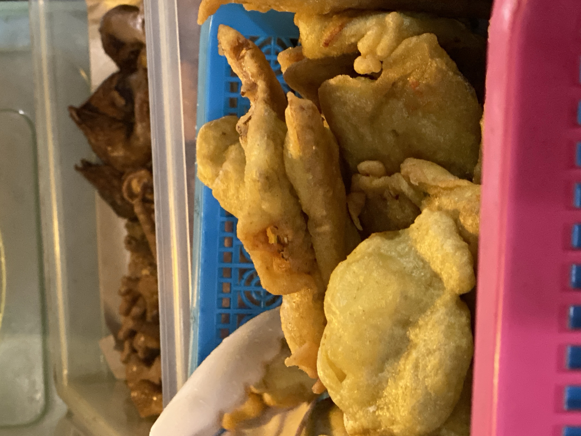 A close-up shot focuses on a basket of fried food, likely at a street food stall in Kasihan, Indonesia. The fried items, predominantly golden-yellow and appearing somewhat irregular in shape, are piled high in a blue plastic basket. Some pieces have a lighter, doughy appearance, while others are darker and crispier. In the background, other food items are visible in containers, including what appears to be some darker, cooked pieces. A white utensil is partially visible, suggesting it's used for serving. The foreground is dominated by the bright pink rim of another container, creating a strong color contrast with the food. The lighting suggests an indoor or covered outdoor setting, common for food vendors.