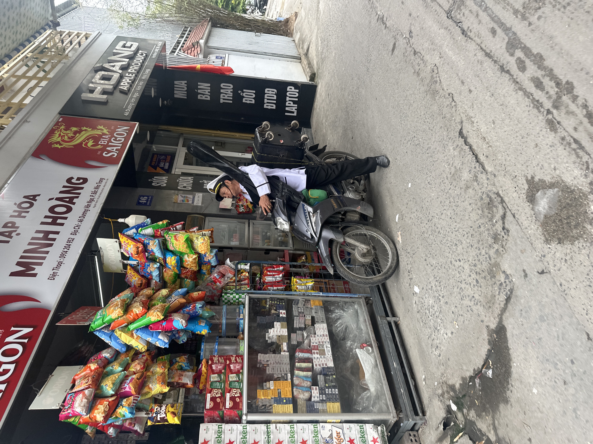 A man in a white shirt and sailor hat is laying on a gray motorcycle parked on the sidewalk in front of a store with Vietnamese signs in Bac Nha Trang Ward, Vietnam. A black suitcase on wheels and a long, black case are strapped to the back of the motorcycle. The store's signage includes "MINH HOANG" and "SAIGON" and indicates the address as "46 Dương Vân Nga P. Bắc Nha Trang". A second sign reads "HOANG APPLE PRODUCT 46 Dương Vân Nga - Vĩnh Hải - Nha Trang". The store is stocked with snacks, cigarettes, and beer, including Heineken and Tiger. The street appears to be paved and somewhat dirty.
