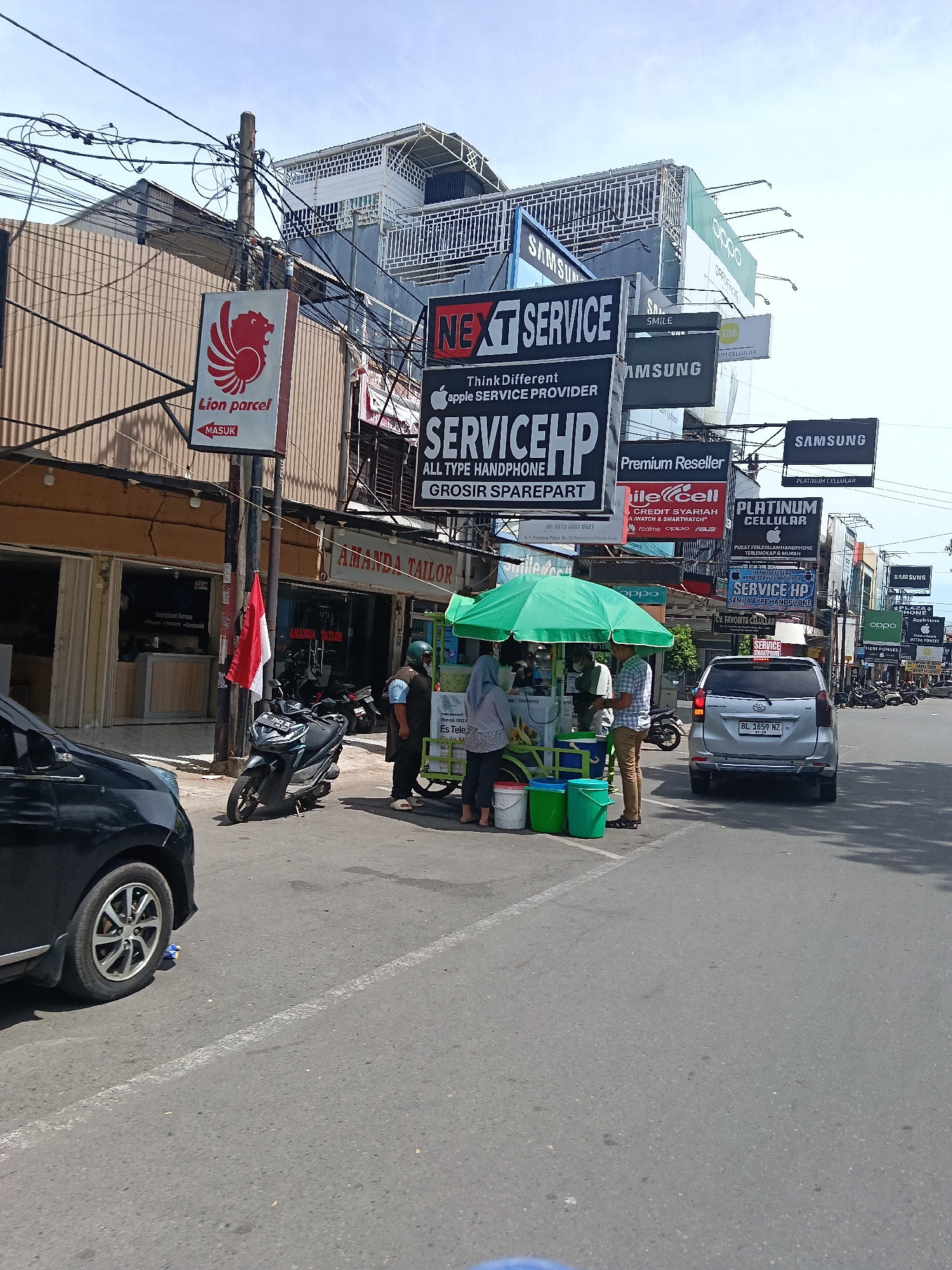 The image shows a street scene in Banda Aceh, Indonesia, on a sunny day. A silver Toyota Avanza with license plate BL 1859 NZ is parked on the right side of the road. Several people are gathered around a green umbrella-covered cart that appears to be selling drinks or food. A black sedan is on the left side of the road. Various signs are visible overhead, including those for "Lion Parcel," "Next Service," "Service HP," and "Samsung." A motorcycle is parked near a pole with a red and white Indonesian flag. The area appears to be a commercial district with multiple shops and businesses. The road is asphalt, and there are utility poles with a multitude of wires crisscrossing overhead.