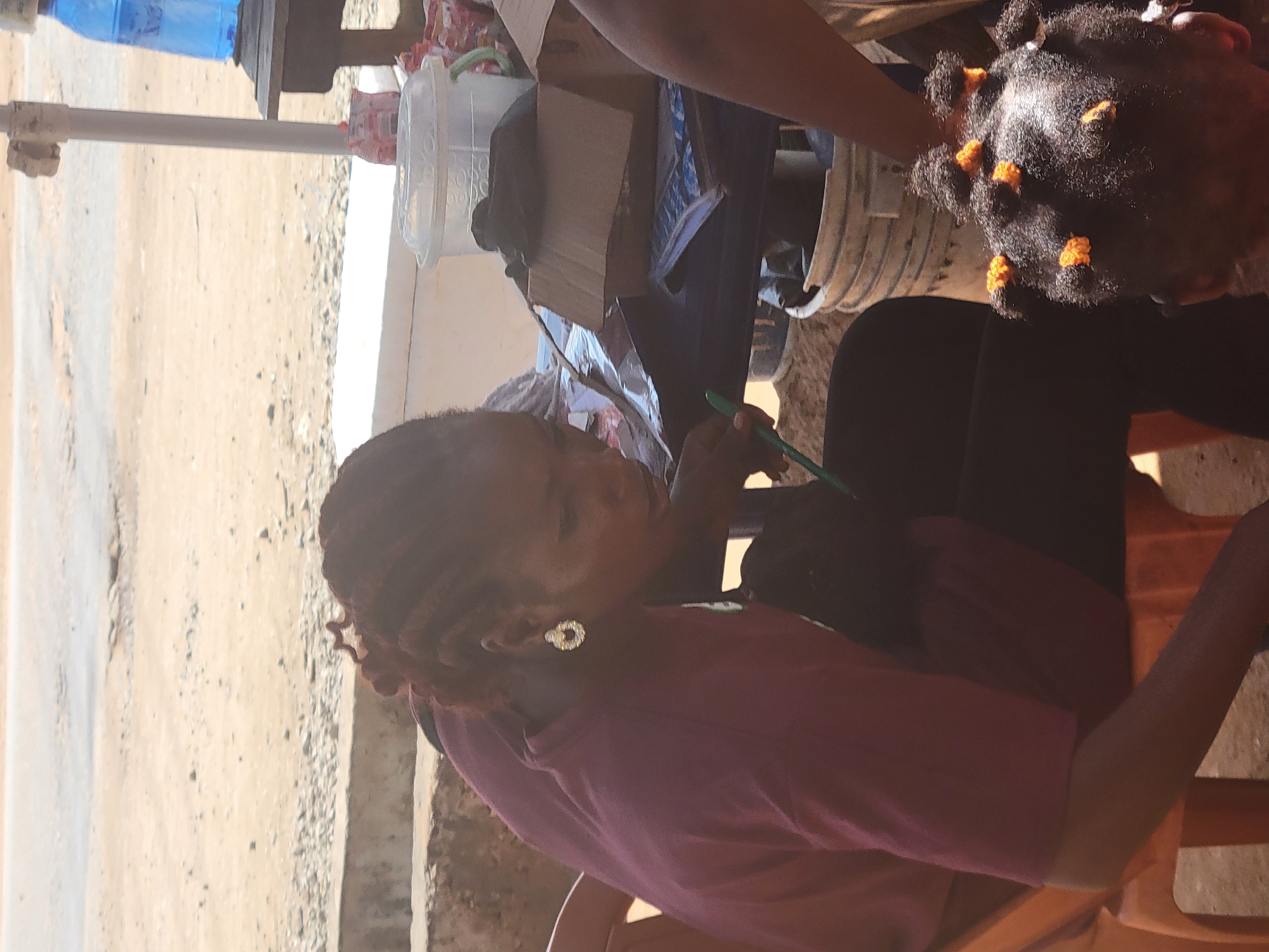 The image depicts an outdoor scene in Kwamba, Nigeria, under bright daylight. In the foreground, a woman with braided hair and large hoop earrings is seated at a table. She is wearing a maroon-colored t-shirt and appears to be looking down, possibly engaged in an activity on the table. Her left arm rests on her lap. To her right and slightly behind her, a young child with braided hair adorned with orange beads is visible from the back, also looking down.
The table is cluttered with various objects, including a blue notebook, a cardboard box, and a clear plastic container with a green lid. Behind the table, a large blue water jug is visible on a wooden stand, and in the background, a dusty, unpaved area with some rocks and vegetation suggests the natural environment of the location. The overall atmosphere appears casual, possibly a market stall or a roadside setup.