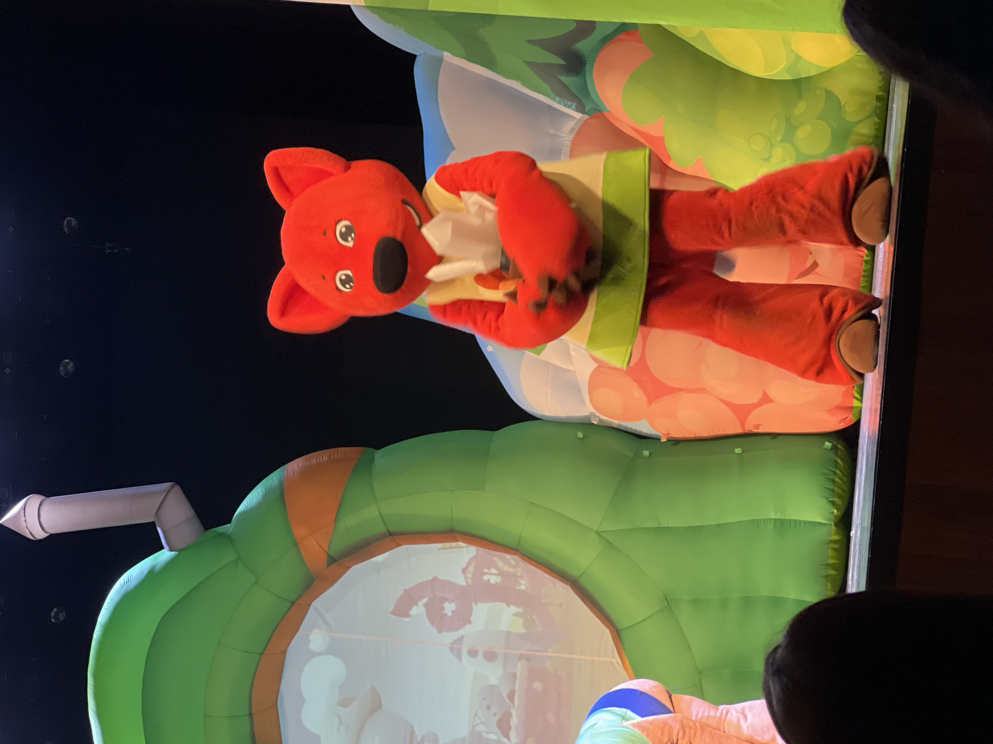 The image shows a stage performance or a public appearance of a mascot character that resembles a red-orange animal, possibly a fox or koala. The character is standing inside an inflated structure. The environment surrounding the character includes inflatable elements, with a large green and orange inflated arch. The location is inferred to be in the city of Chaikovsky, Russia. The image appears to be taken during a show or event, potentially in a theater or event space, with an audience member visible in the foreground. The scene is illuminated by stage lights, creating a vibrant and dynamic atmosphere.