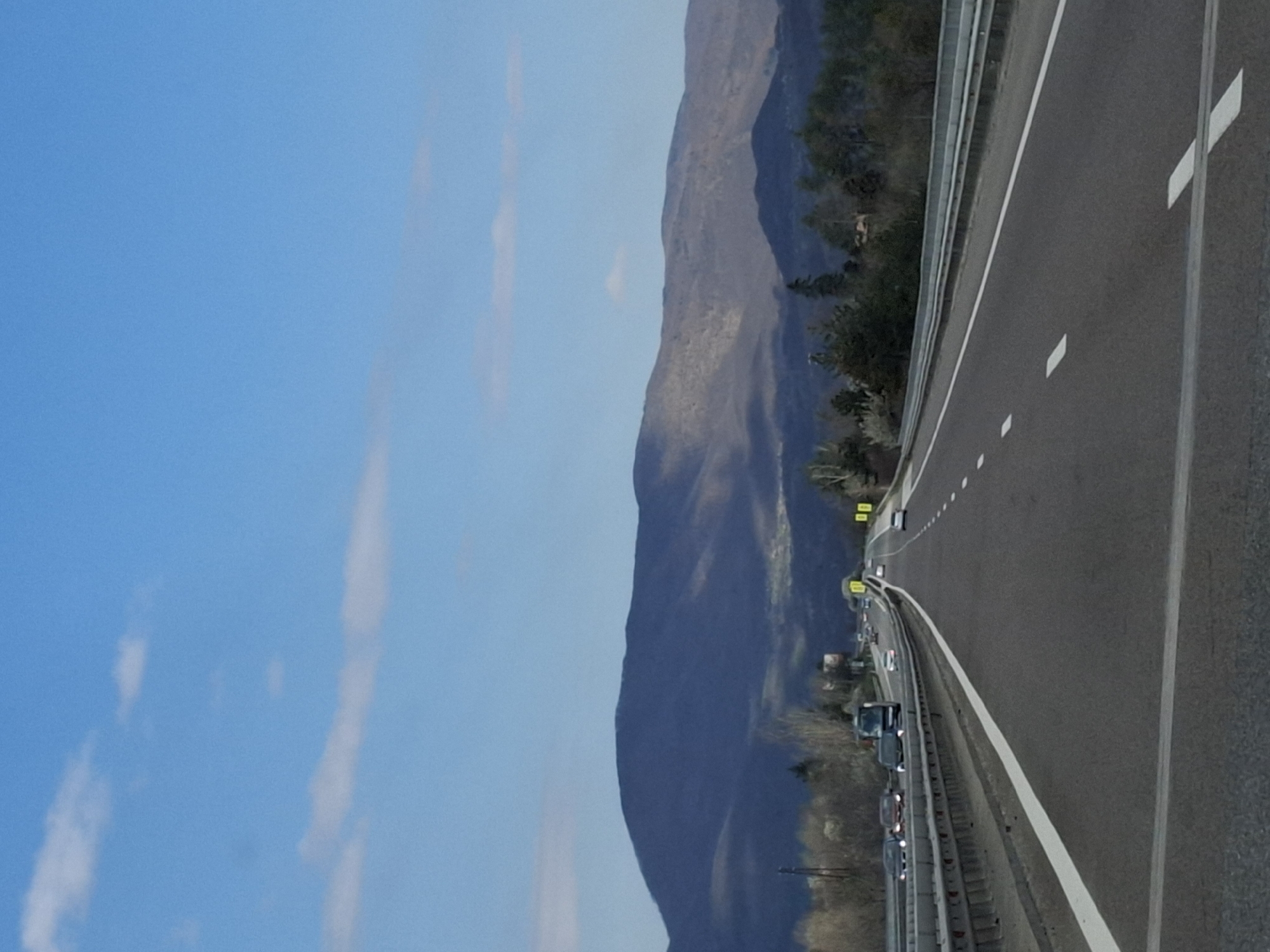 A highway with multiple lanes is visible, lined with a guardrail. Traffic is present, with several vehicles moving along the road. To the left of the highway is a large mountain range. The sky is blue with some streaks of light clouds. The picture may have been taken near Церово, Bulgaria.