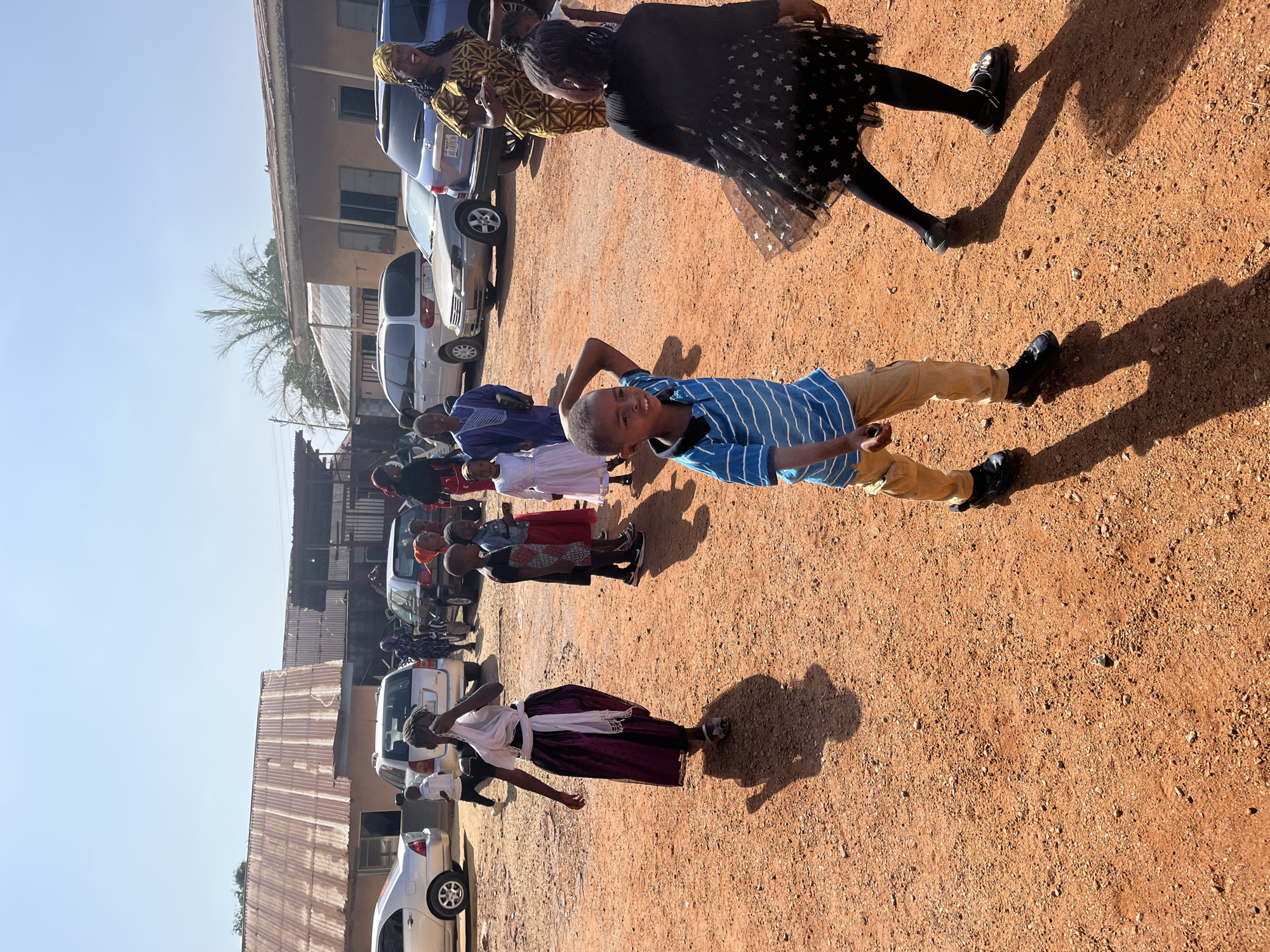 The image depicts a group of people standing outside, presumably in Gura Top, Nigeria. The ground is reddish-brown and gravelly. A young boy wearing a blue and white striped shirt and tan pants is in the foreground. Behind him are several adults and children, some wearing traditional clothing. Several cars are parked nearby. The sky is pale blue, suggesting it is daytime. Buildings with corrugated metal roofs are visible in the background.
