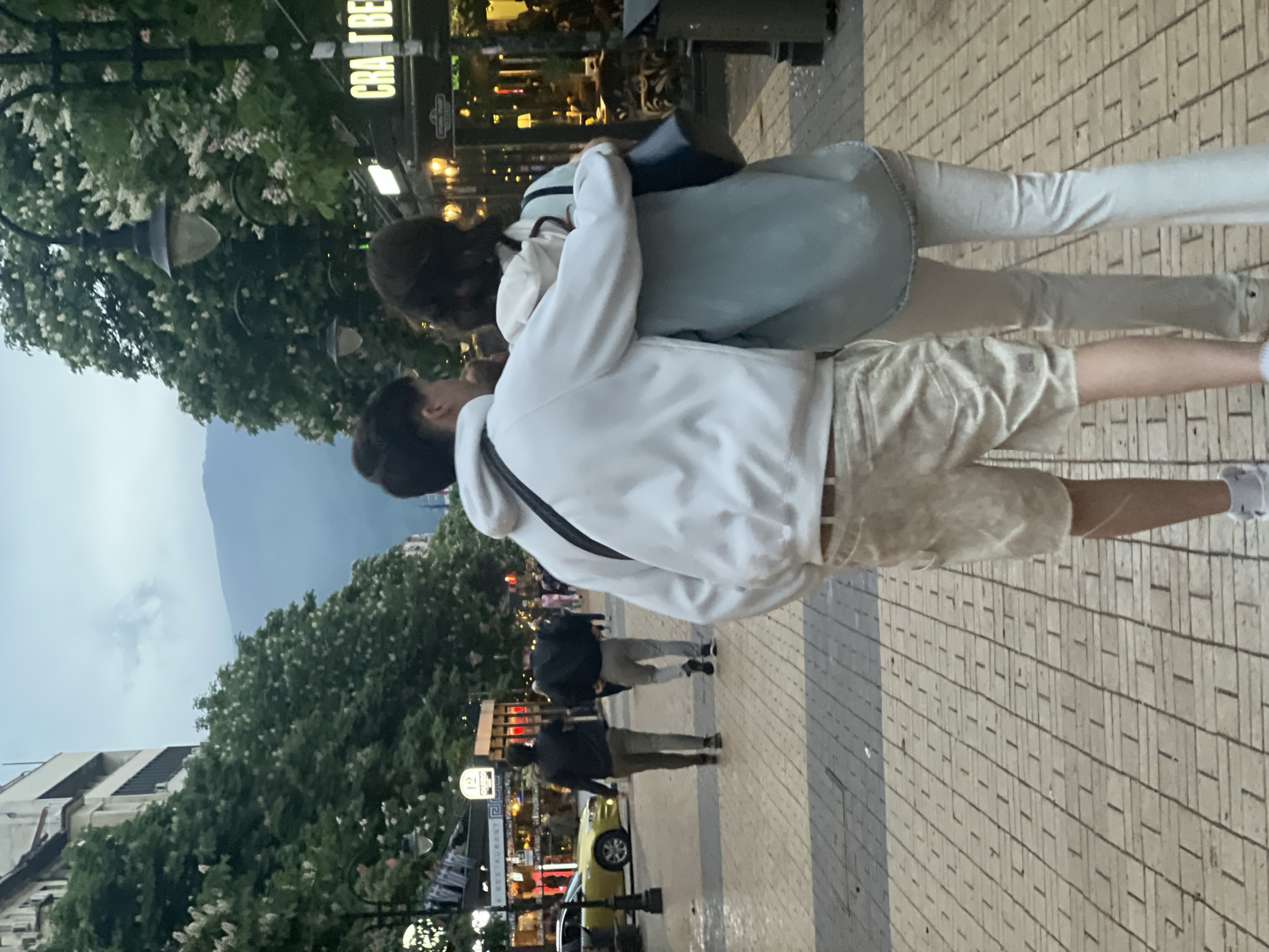 A horizontal, outdoor photograph captures two people embracing on a brick-paved street. A man wearing a white hoodie and light-colored shorts is on the left and an individual in a light-blue jacket and light-colored pants is on the right. Both individuals have dark hair. Their faces are close, suggesting a kiss or embrace. A black strap is visible on the man’s shoulder. The background is a city street with trees, lampposts, and buildings. "CRAFT BEER" is visible on a sign. A mountain can be seen in the distance. Other pedestrians and a light-yellow car are also visible. The image appears to be taken in Sofia, Bulgaria.