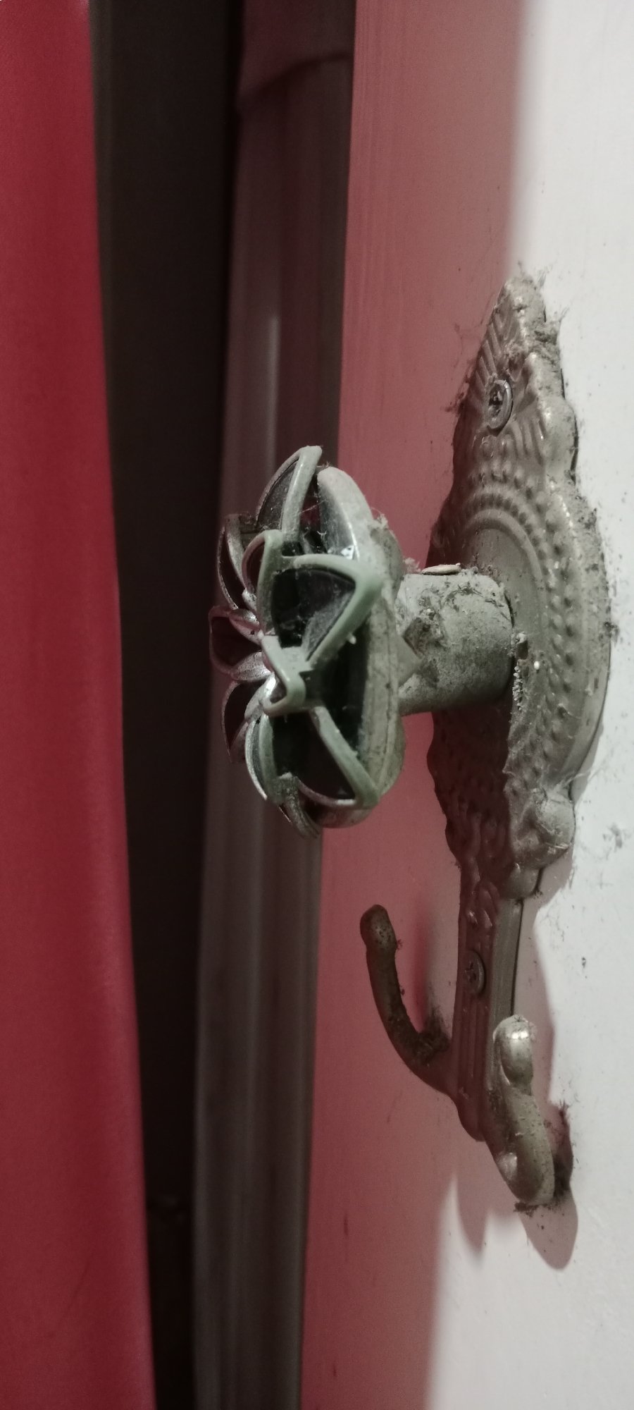 The image shows a dusty, ornate metal hook mounted on a white wall. The hook has a decorative backplate and a single curved arm. Attached to the hook is a small, fan-shaped object, possibly a decorative finial or part of a disassembled item, made of a light green material and metal. The object is covered in dust and cobwebs. To the left of the hook, there are two vertical elements, one deep red and the other dark gray, possibly curtains or drapery. The scene appears to be indoors, in a dimly lit area. There is no indication of Soko, Indonesia, or any specific location within a city.