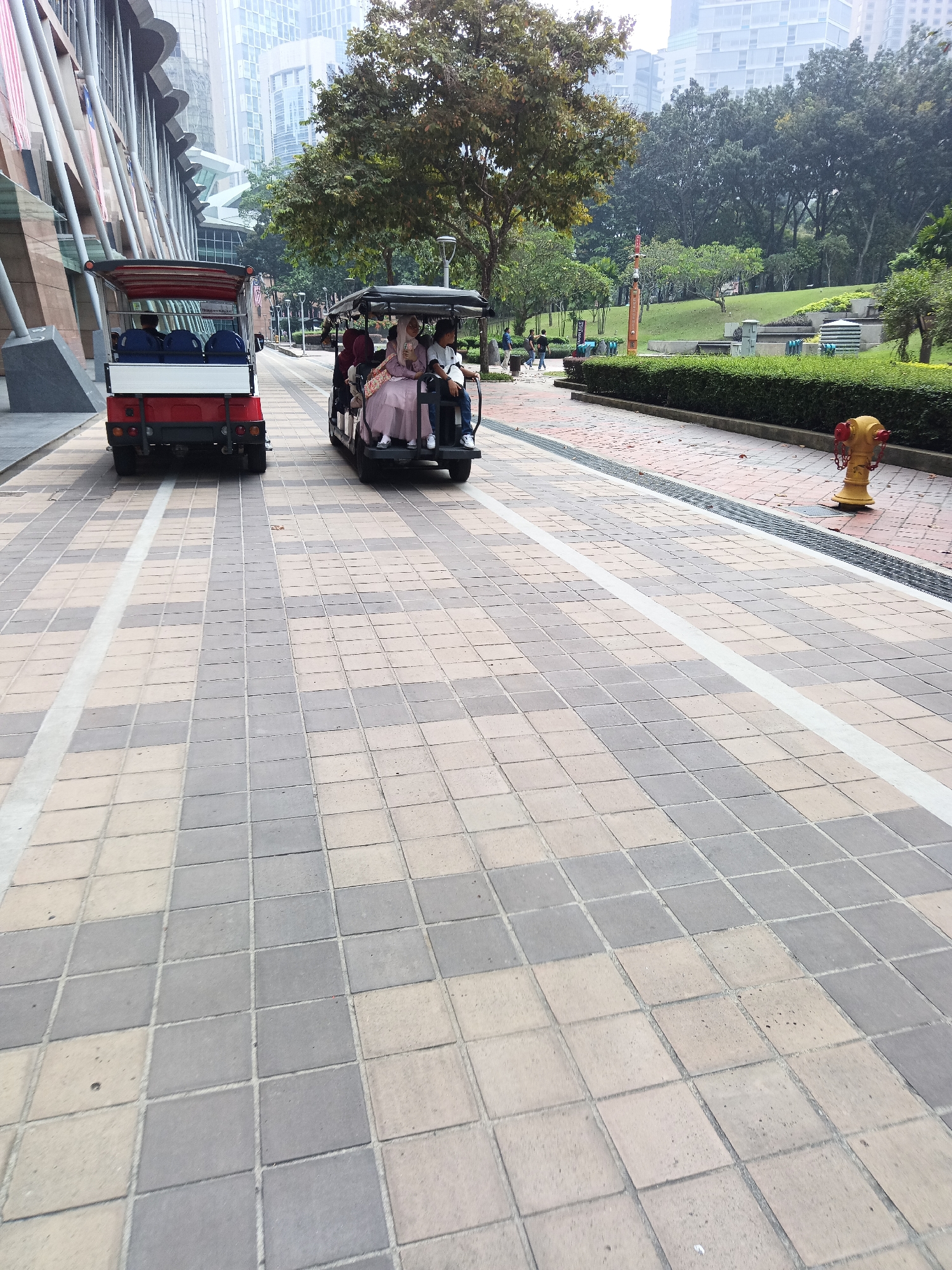 The image depicts a paved walkway in Kuala Lumpur, Malaysia, likely within a park or urban green space adjacent to a modern building. Two electric carts are visible on the walkway. The cart in the foreground is red and white and appears to be empty, while the cart behind it, which is black, carries four people. The people in the black cart include a woman in a pink hijab and a pink dress, a man in a white shirt and blue pants, and two other individuals whose faces are not clearly visible.
The walkway is paved with square tiles in a checkered pattern of beige and gray. To the right of the walkway, there is a manicured green hedge, a yellow fire hydrant, and a sloped grassy area with trees and a water feature in the background. The background also shows modern high-rise buildings and trees, suggesting an urban setting. The atmosphere appears to be overcast or hazy.