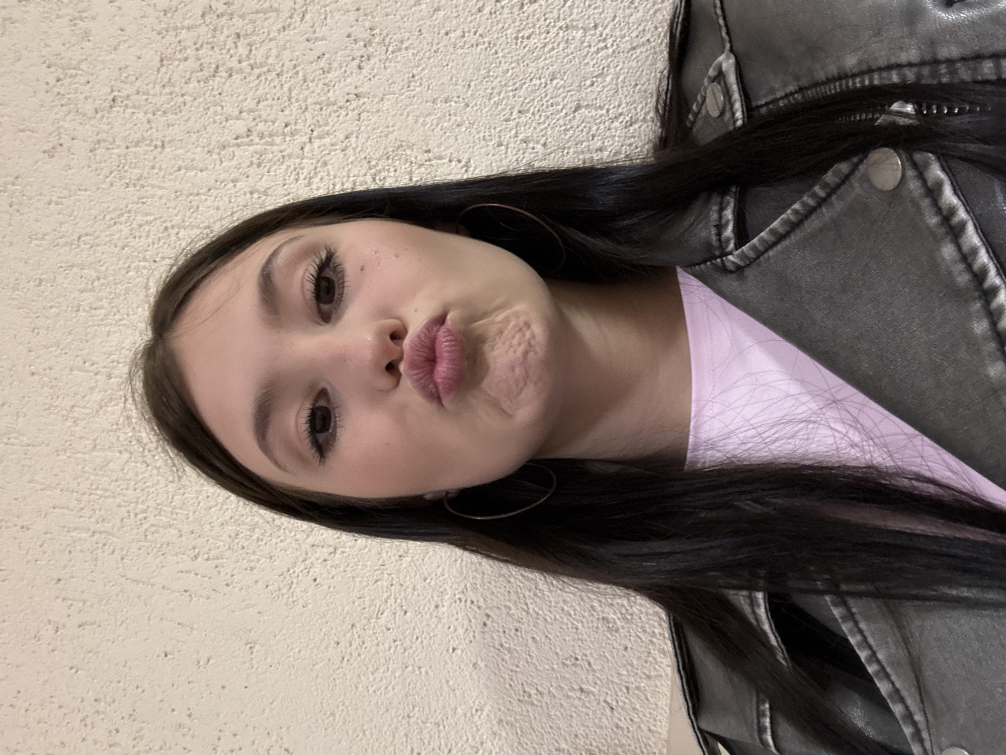 A young woman poses indoors against a textured, off-white wall, likely within a building in Chaikovsky, Russia. She has dark hair, fair skin, and brown eyes, and is making a "kissing" face at the camera. She wears hoop earrings, a light pink shirt, and a distressed gray leather jacket. The top right of the image reveals the jacket's stitching and a metal snap. The location is implied through context.