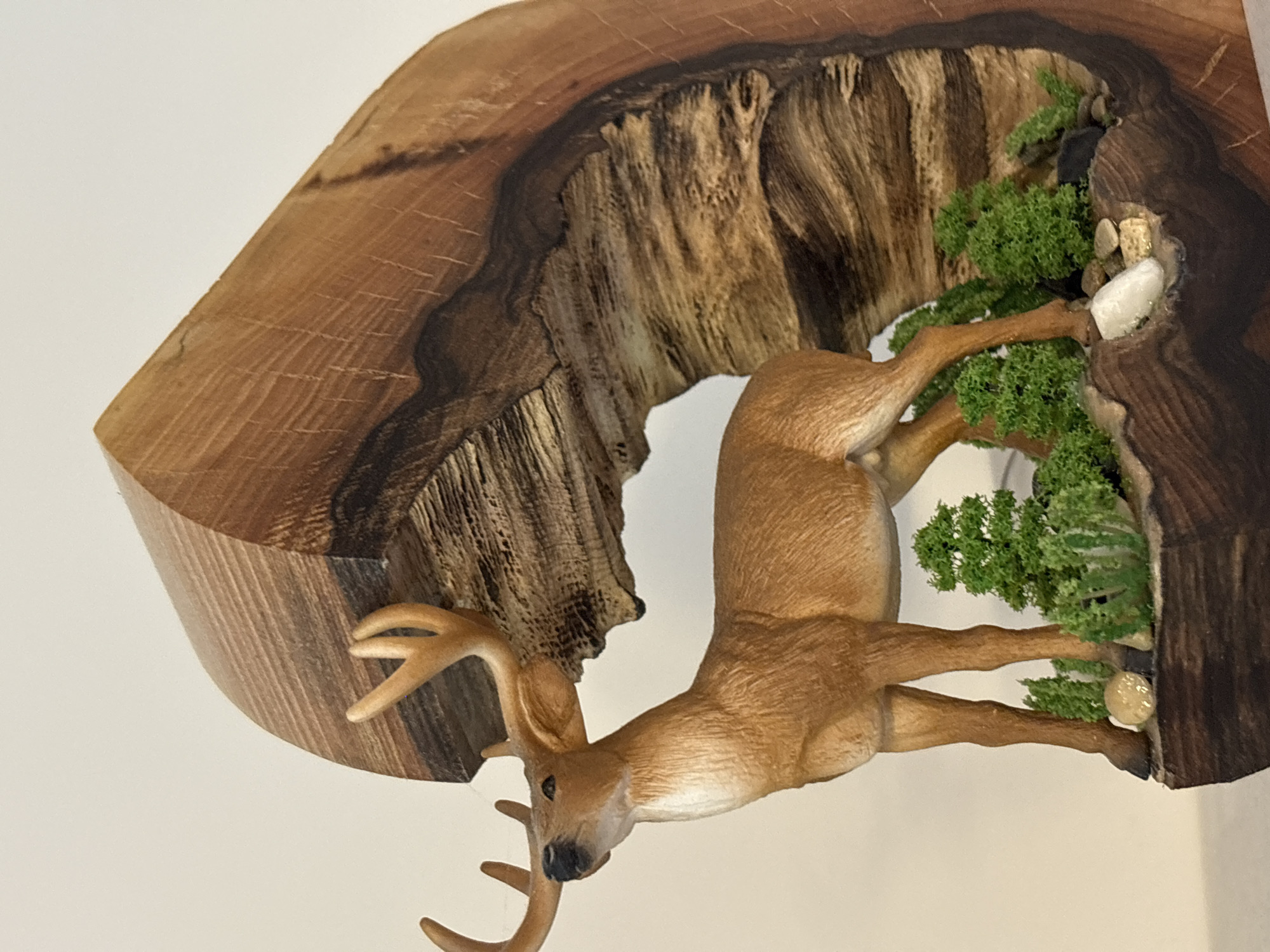 The image shows a diorama of a deer standing in a naturalistic setting. The deer is a brown figurine with antlers, positioned on a wooden base. Behind the deer is a carved wooden structure that resembles a hollowed-out log or a cave opening. The base of the diorama is decorated with small green artificial trees and scattered stones. The background is a plain, light-colored wall, indicating the diorama is likely displayed indoors. There is no visible indication of the city of Plovdiv, Bulgaria in the image.