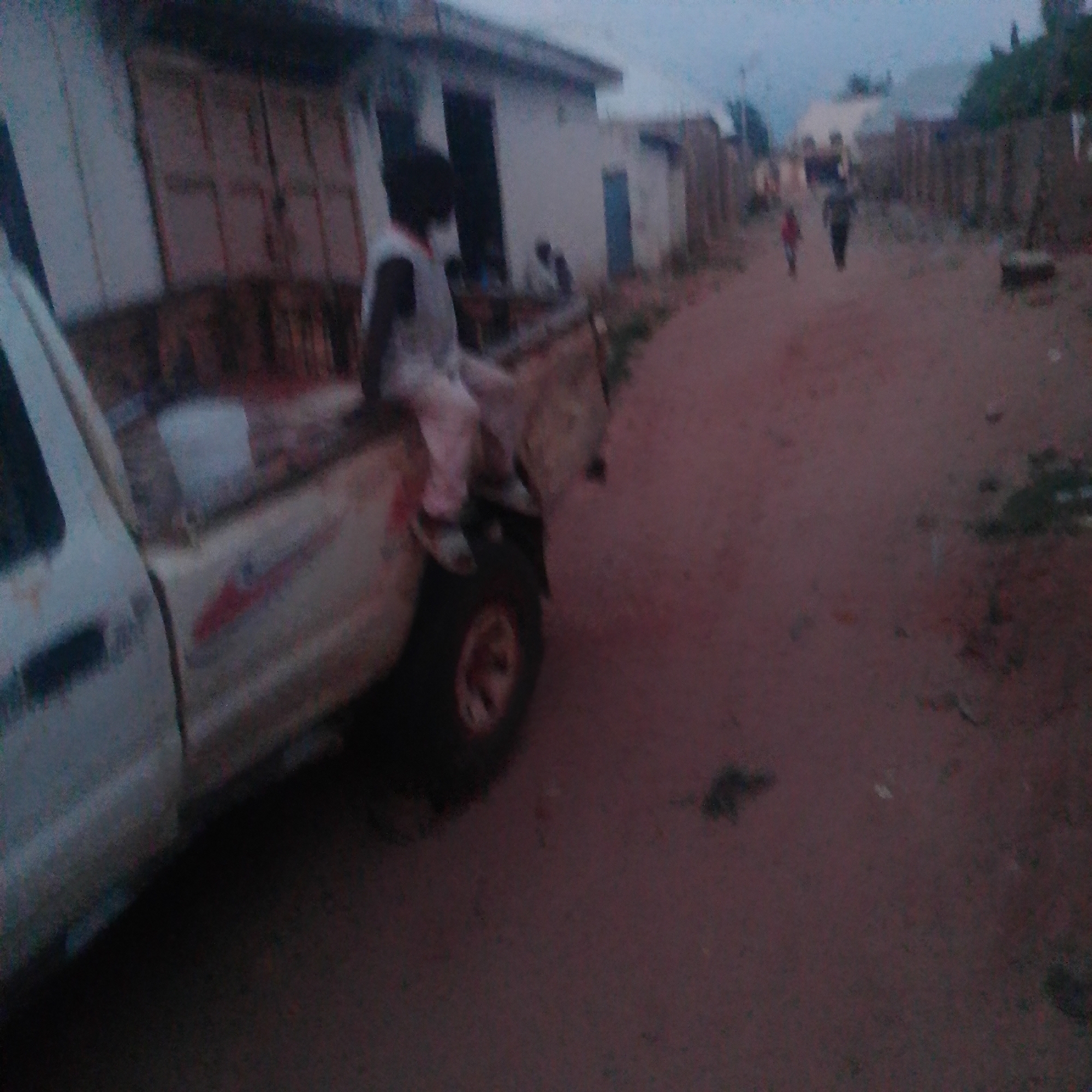 In Gombe, Nigeria, on a dirt road with buildings lining one side, a person is seated on the tailgate of a white pickup truck. The individual is wearing a light-colored shirt and pants. The truck's bed appears to be empty. In the background, a few other individuals are visible walking down the road, and some structures are present. The environment suggests a somewhat rural or less developed urban setting, with red soil and a cloudy sky.