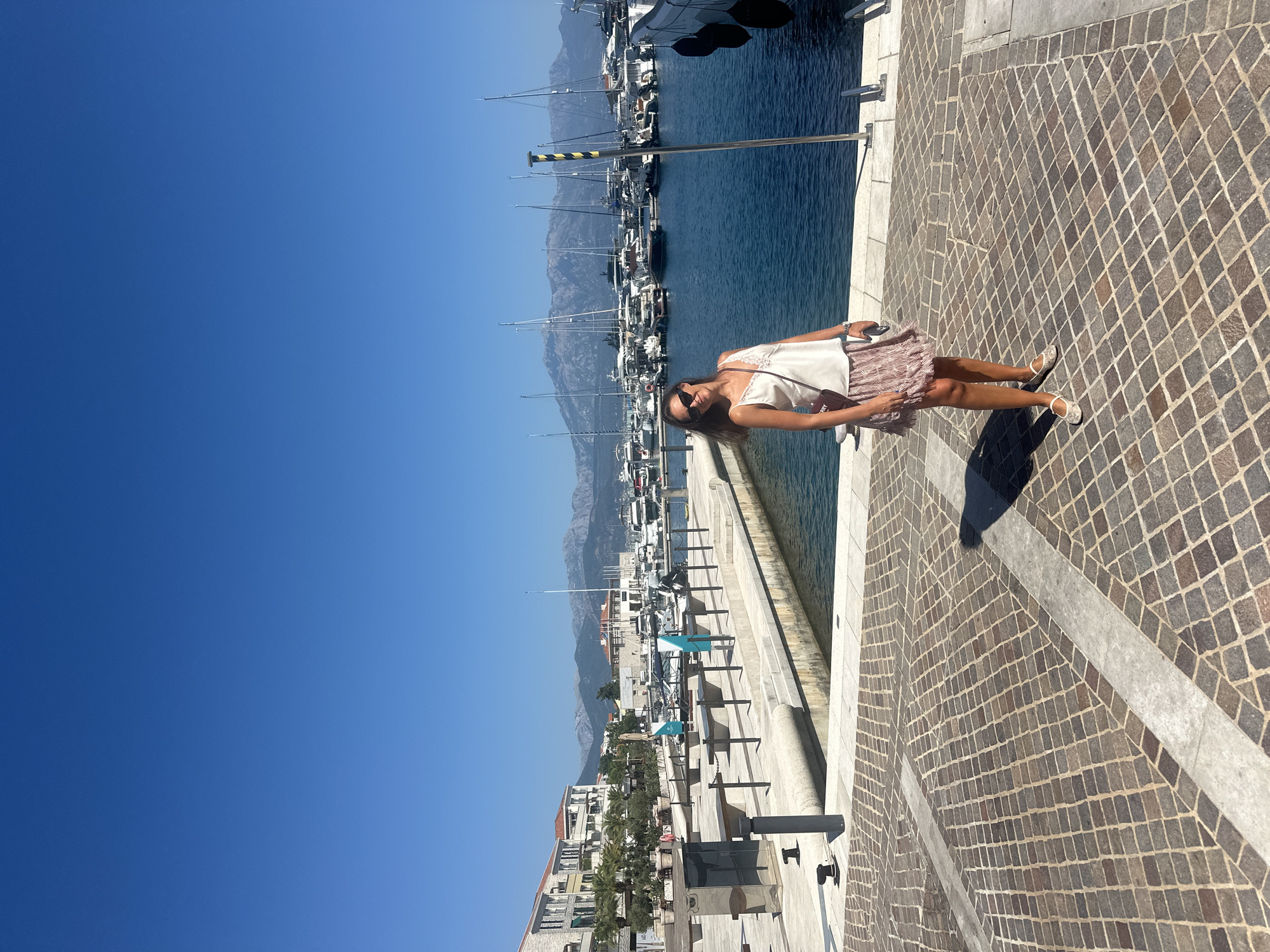 A woman stands on a stone-paved promenade in the city of Herceg Novi, Montenegro, with a marina full of yachts and sailboats behind her. The sky is clear and blue, and the water in the marina is calm and dark. The woman is wearing a white camisole, a patterned skirt, and white flats. She has sunglasses on and a purse slung over her shoulder. In the background, there are buildings with red roofs, palm trees, and mountains in the distance. The promenade is lined with bollards and benches. The scene appears to be a sunny day in a coastal city.