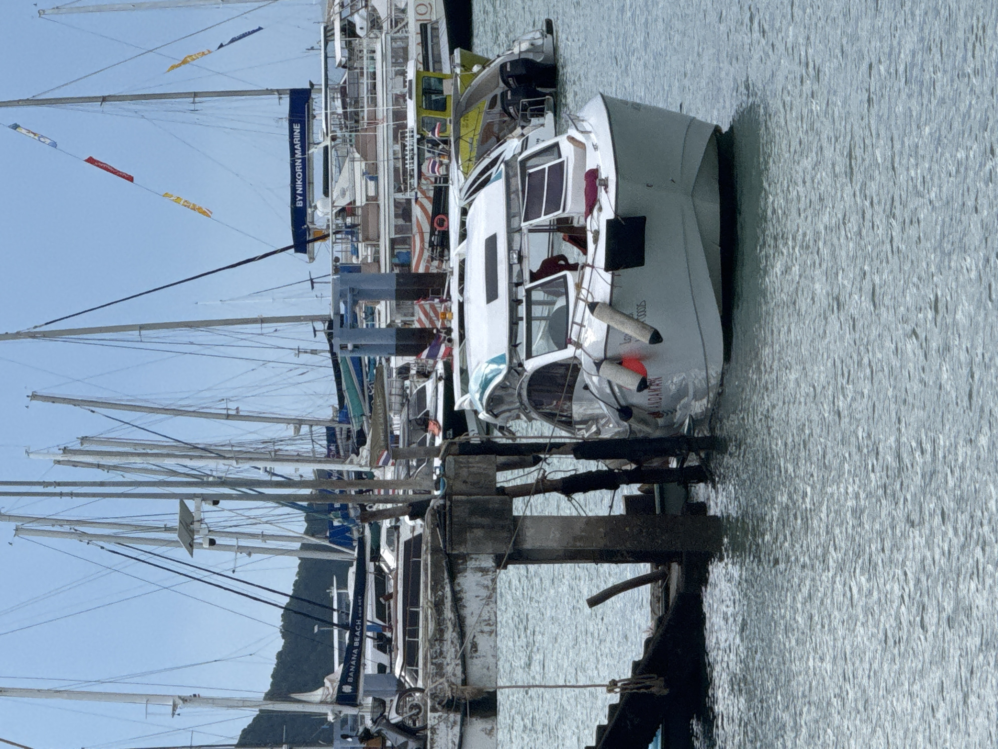 The image shows a harbor in Chalong, Thailand, with numerous boats docked. In the foreground, a white motorboat with blue and green accents is partially obscured by a weathered wooden pier. The boat appears to be listing slightly to the port side, with its bow pointed towards the viewer. The words "ANDAMAN" are visible on the side of the boat. Several buoys are attached to the boat's hull. Behind this boat, other vessels, including sailboats and larger passenger boats, are moored. The water is choppy, reflecting the overcast sky. The pier consists of concrete pillars and wooden planks. In the background, a hillside covered in lush vegetation is visible. The overall scene depicts a busy harbor setting.