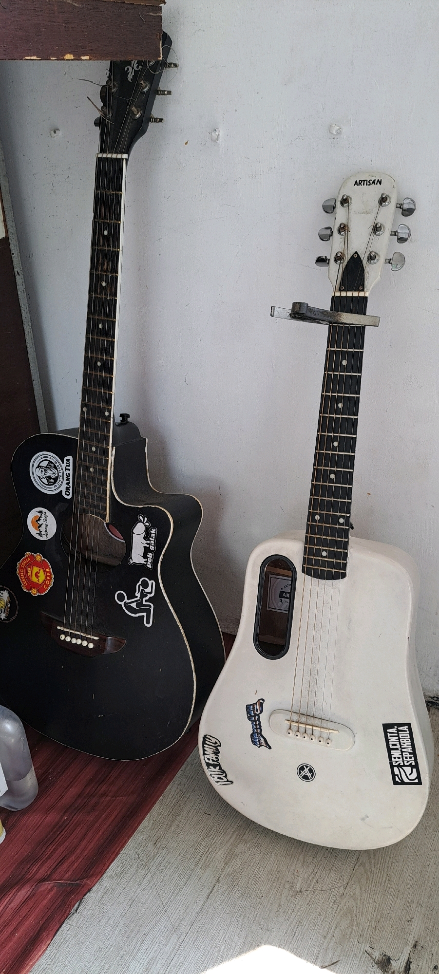 Two acoustic guitars are leaning against a white wall in what appears to be an outdoor or semi-outdoor area with a wooden floor. The guitar on the left is black and adorned with several stickers, including one with the text "ORANG TUA" and another depicting a stylized figure. It has a wooden fretboard and tuning pegs. The guitar on the right is white and has a different shape, featuring an oval soundhole. This guitar also has stickers, one with the text "SENI.CINTA. SEPAKROLA". A capo is attached to the neck of the white guitar. The floor shows a transition from a reddish-brown area to a lighter greyish-brown area. The overall environment suggests a casual setting, possibly a porch or a workshop in Prigen, Indonesia.