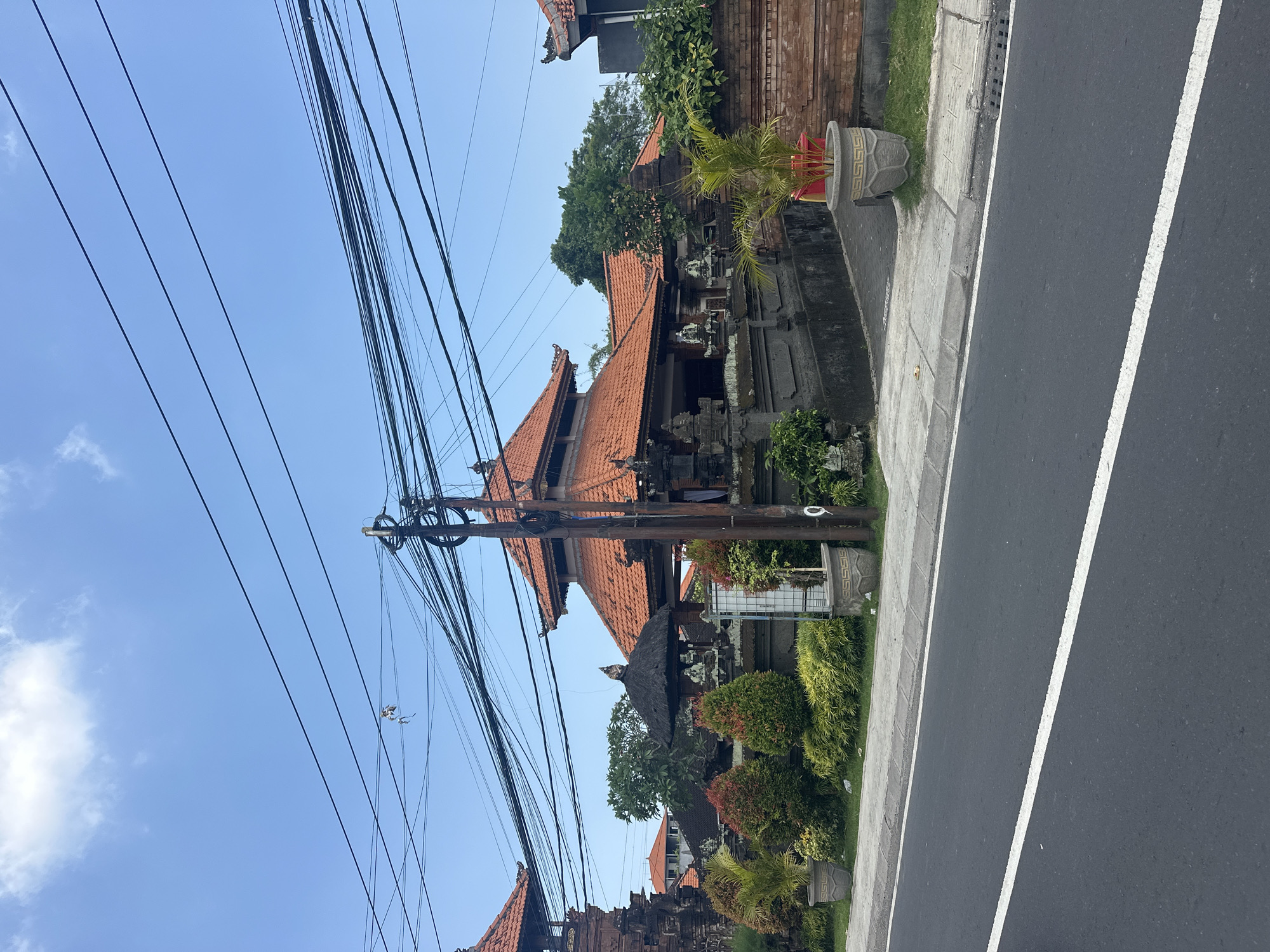 The scene depicts a street view in Dalung, Indonesia, under a clear blue sky with sparse white clouds. In the foreground, a dark asphalt road with a solid white center line extends horizontally. A concrete sidewalk with a raised curb borders the road on the right.
Immediately behind the sidewalk, a property features traditional Balinese architecture. Multiple structures with prominent multi-tiered orange terracotta tiled roofs are visible, constructed from dark grey stone with intricate carvings and decorative elements. A significant utility pole, heavily strung with numerous black electrical and communication wires, stands centrally in the mid-ground, slightly right of the frame's vertical center. A small, light-colored piece of debris is entangled in the overhead wires in the upper left quadrant.
The property boundary is defined by dark stone walls and lush, manicured landscaping. This includes various green shrubs, yellow-green ground cover, and conical bushes with reddish and green foliage. Several decorative grey concrete planters, patterned with a Greek key design, are placed along the sidewalk and within the property, containing small palm-like plants. One building section features a black thatched roof. Further behind the main structures, green trees are visible, and additional red-tiled roofs suggest more buildings in the background. No individuals, vehicles, or specific actions are discernible within the frame.