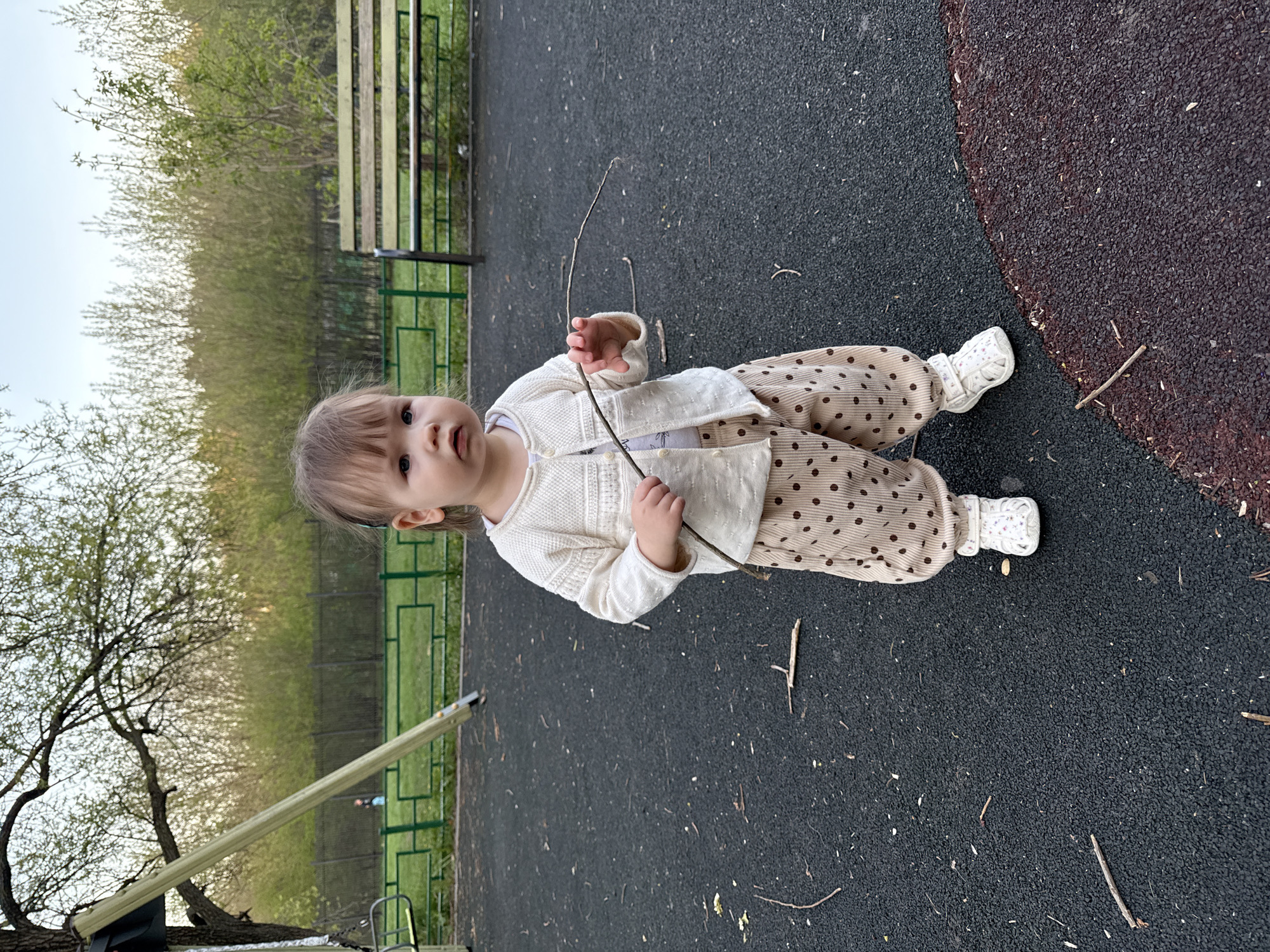 A young child stands on a playground surface that is predominantly black with a circular section of a darker, reddish-brown color. The child is wearing a white cardigan, a light-colored shirt, beige pants with brown polka dots, and white shoes. The child holds a small, thin branch in both hands. Several other twigs and small debris are scattered on the ground. In the background, there is a green metal fence and a wooden bench. Trees and other vegetation are visible beyond the fence. The location is a park or playground in Moscow, Russia.