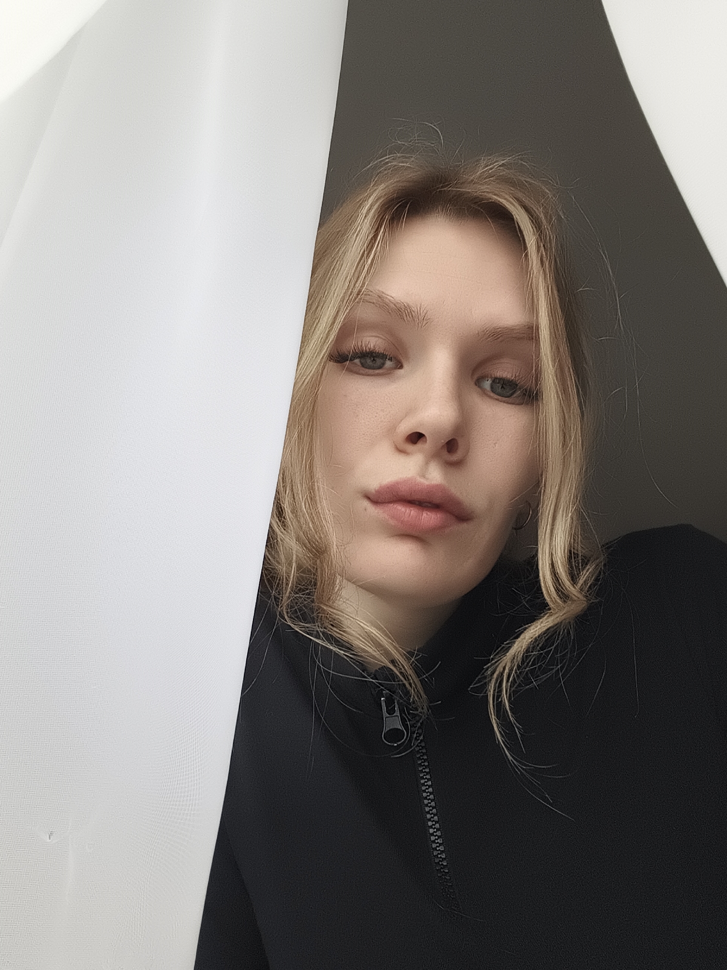 A young, fair-skinned woman with blonde hair is depicted in a close-up shot. She is wearing a black zippered garment. A white curtain is partially obscuring the left side of the frame. The background to the right of the woman is a dark, undefined space. The location is unknown, as is the date the photograph was taken. There is no information to ascertain the location as Русский Вожой, Russia.
