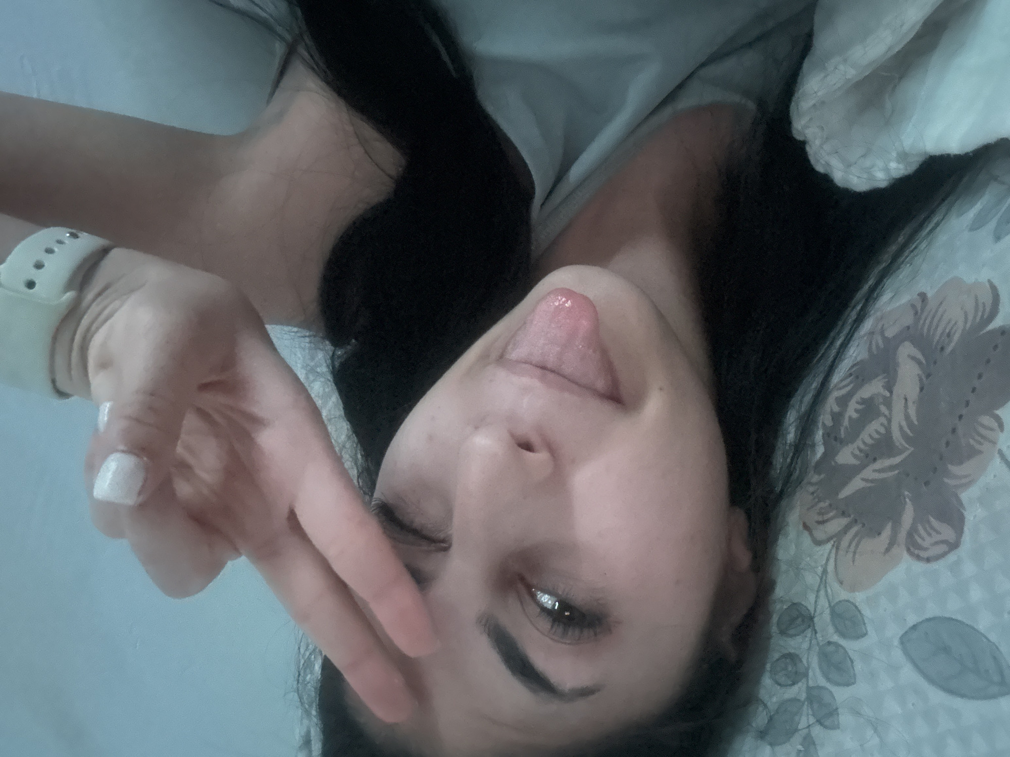 A person with dark hair is lying down on a bed with a floral patterned sheet. The person is sticking their tongue out and making a peace sign with their hand. A watch is visible on their wrist. The person's eye is partially closed. There is no information to confirm this image was taken in the city of Devin, Bulgaria.